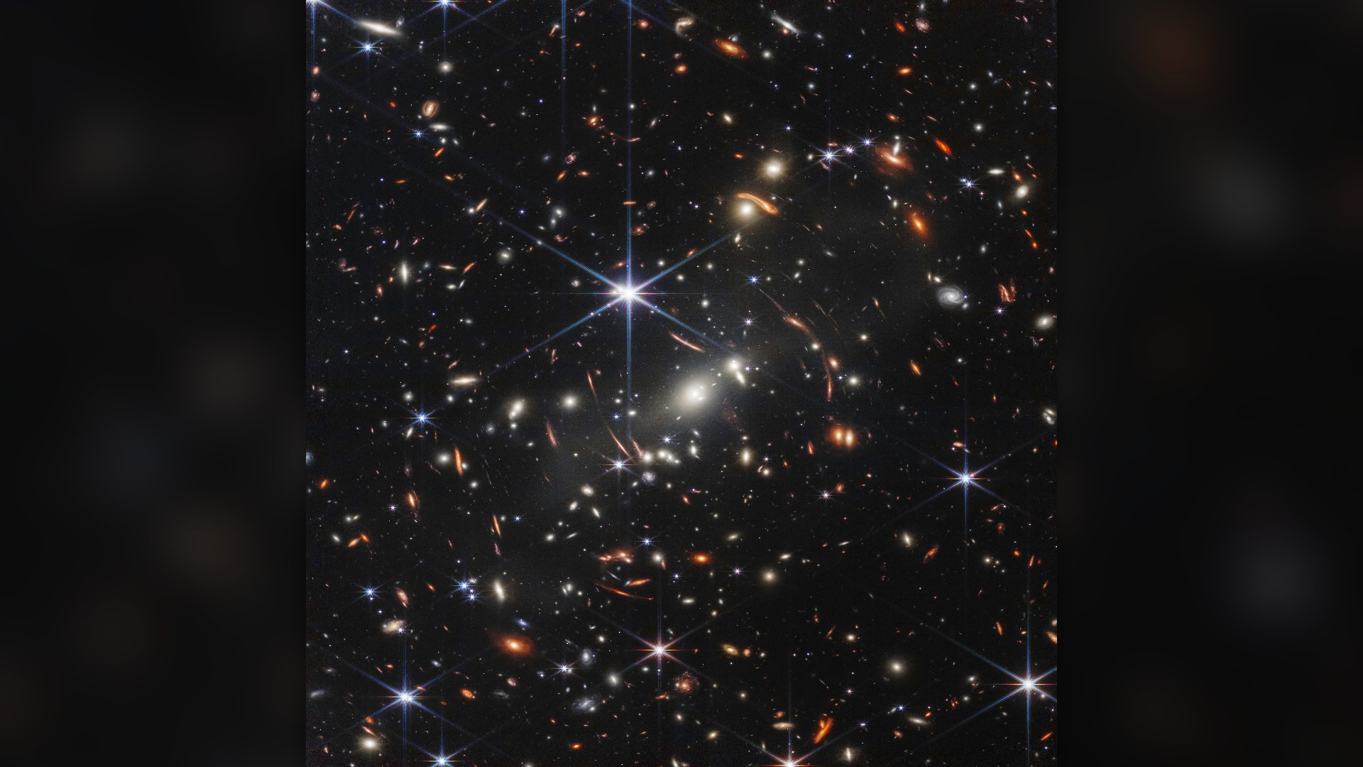 Webb's First Deep Field showing thousands of galaxies in the deep universe around massive cluster SMACS 0723, with distinct orange and red gravitationally lensed arcs.