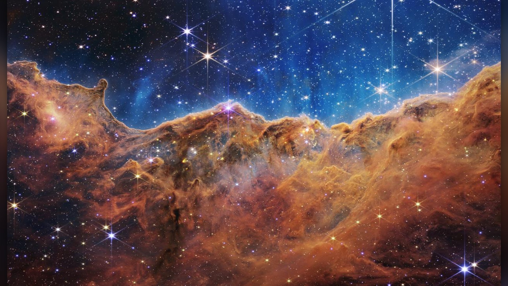 James Webb Space Telescope near-infrared view of the Cosmic Cliffs in Carina, featuring jagged golden-brown dust ridges against a deep blue starry sky.