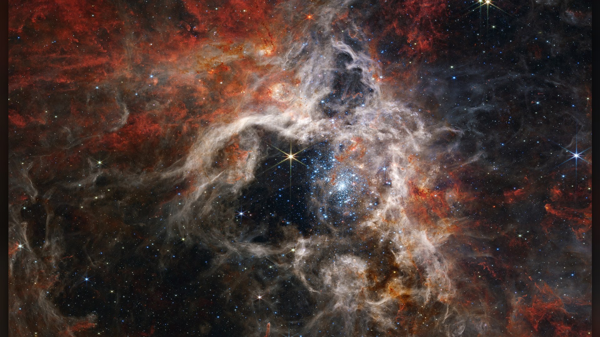 James Webb Space Telescope composite infrared image of the Tarantula Nebula, showing blue newborn star clusters and web-like filaments of orange and tan dust.