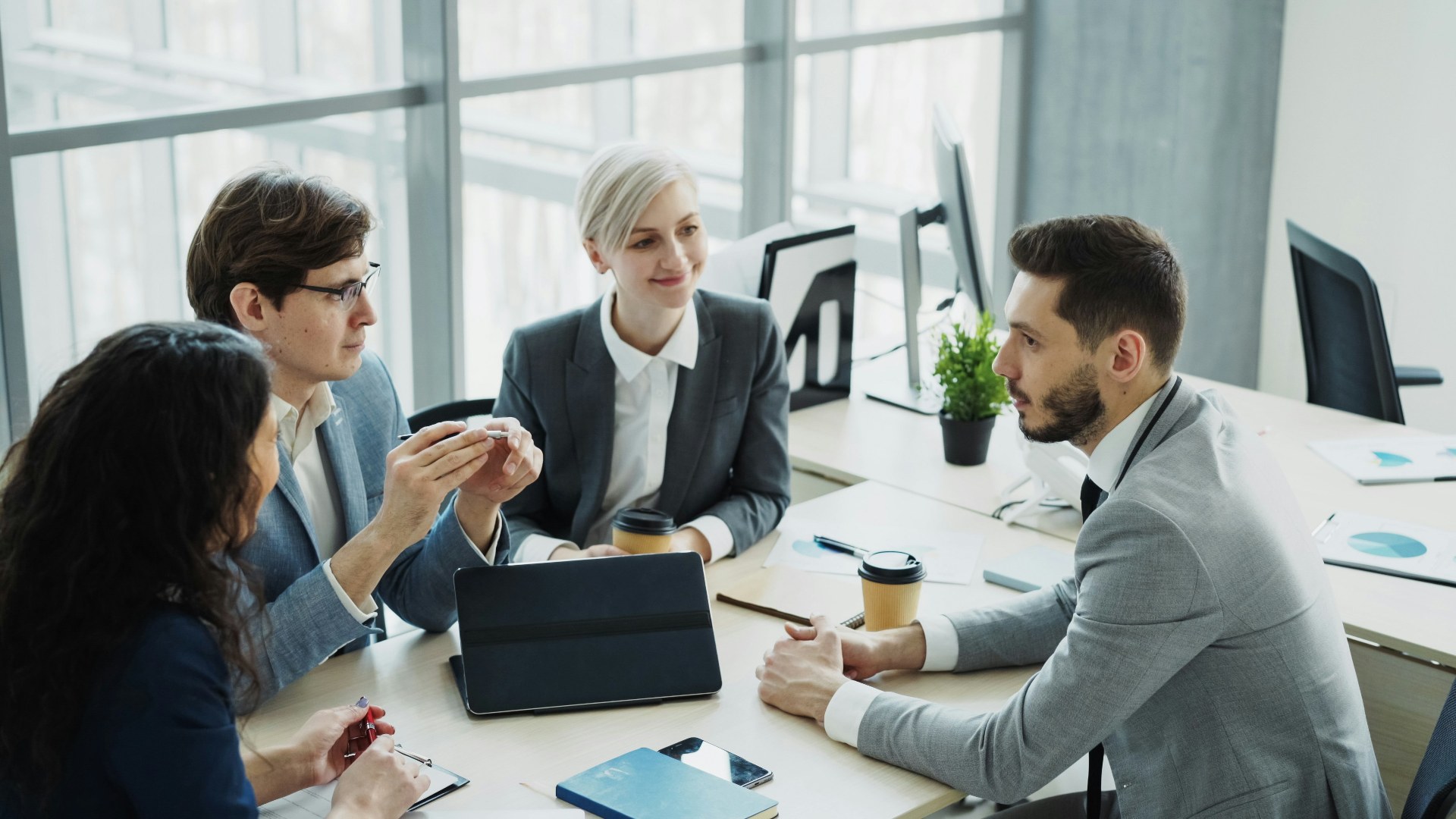 Diverse team of corporate coworkers talking together around laptops in a modern office.