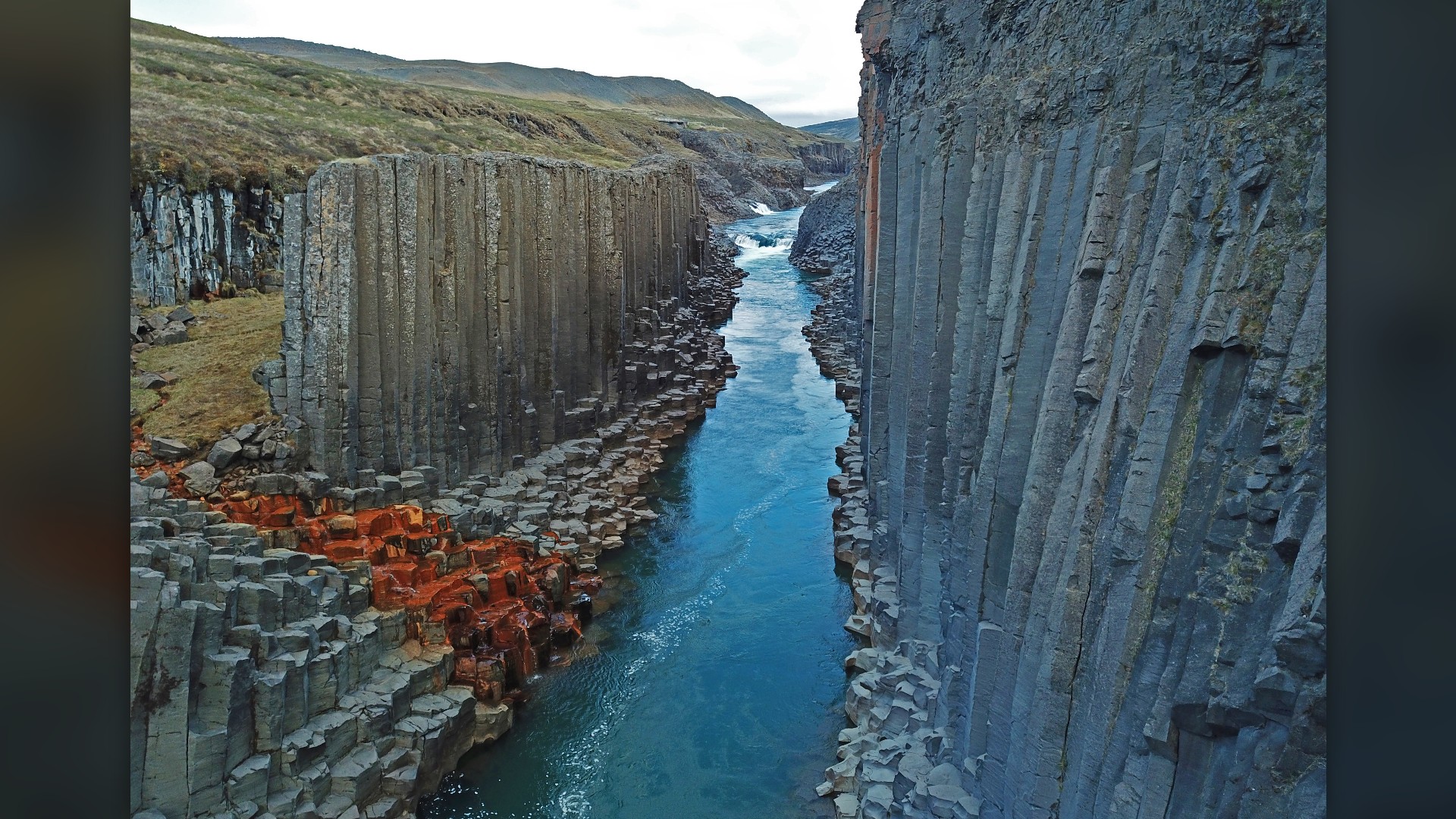 Turquoise river water flowing between geometric hexagonal basalt columns in Stuðlagil Canyon, Iceland.