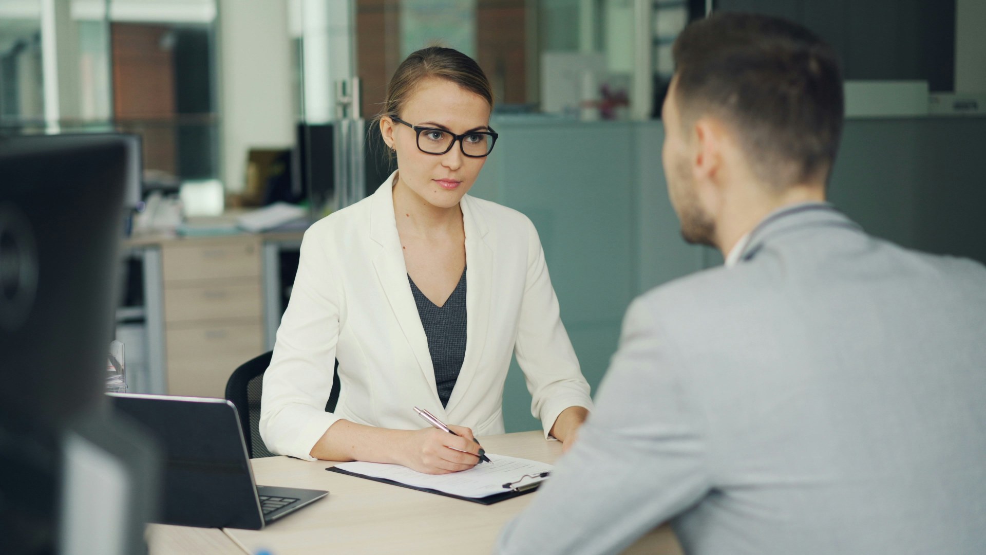 Corporate manager in glasses interviewing an employee across an office desk.