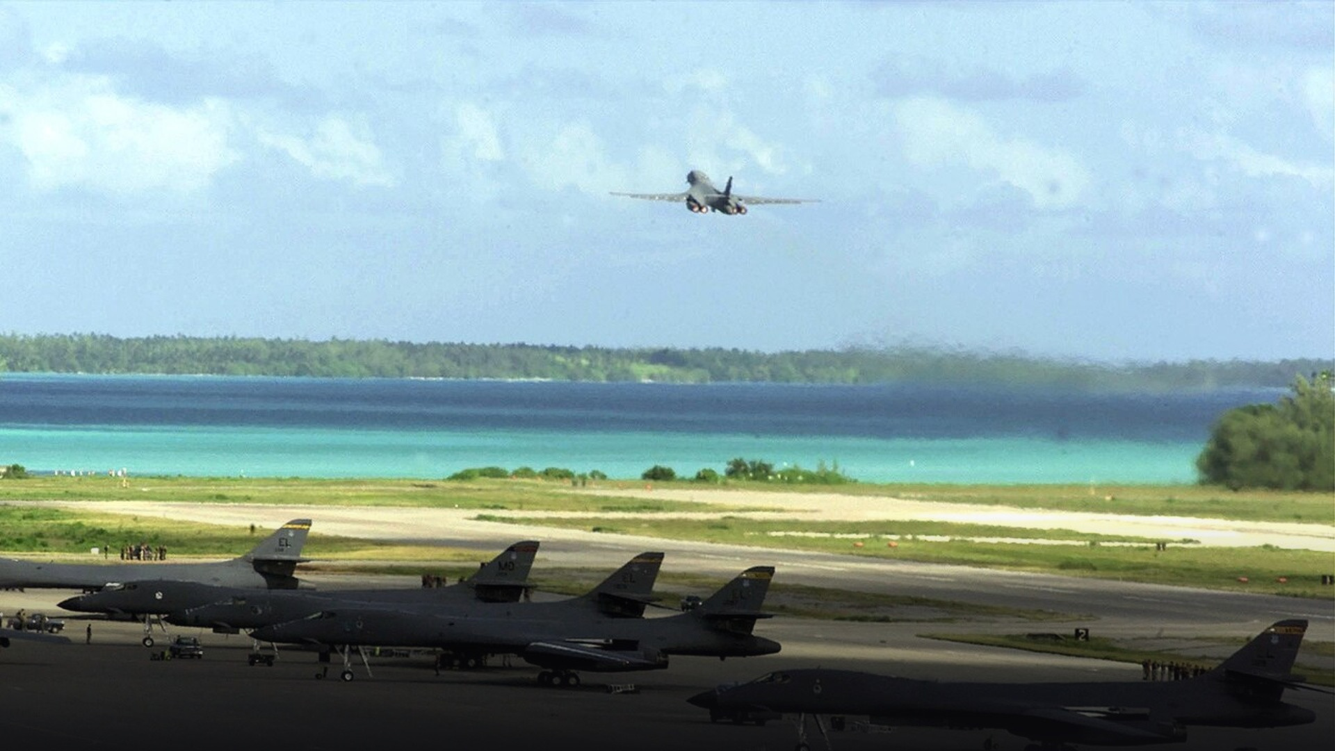 U.S. Air Force B-1B Lancer supersonic strategic bombers parked on the Diego Garcia flightline with another launching over the tropical lagoon.