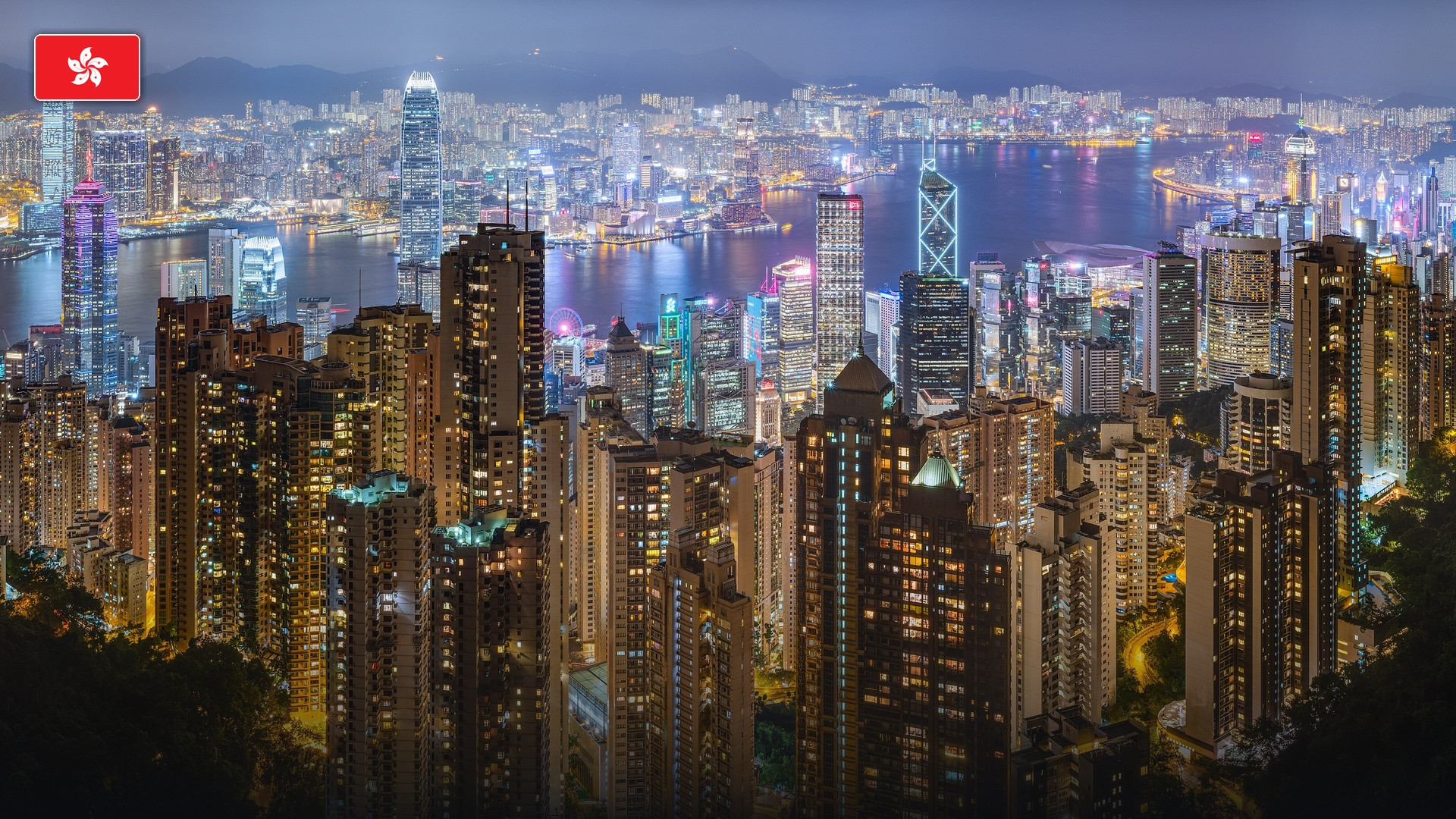 The illuminated skyscrapers of Victoria Harbour viewed from Victoria Peak, with the flag of Hong Kong.