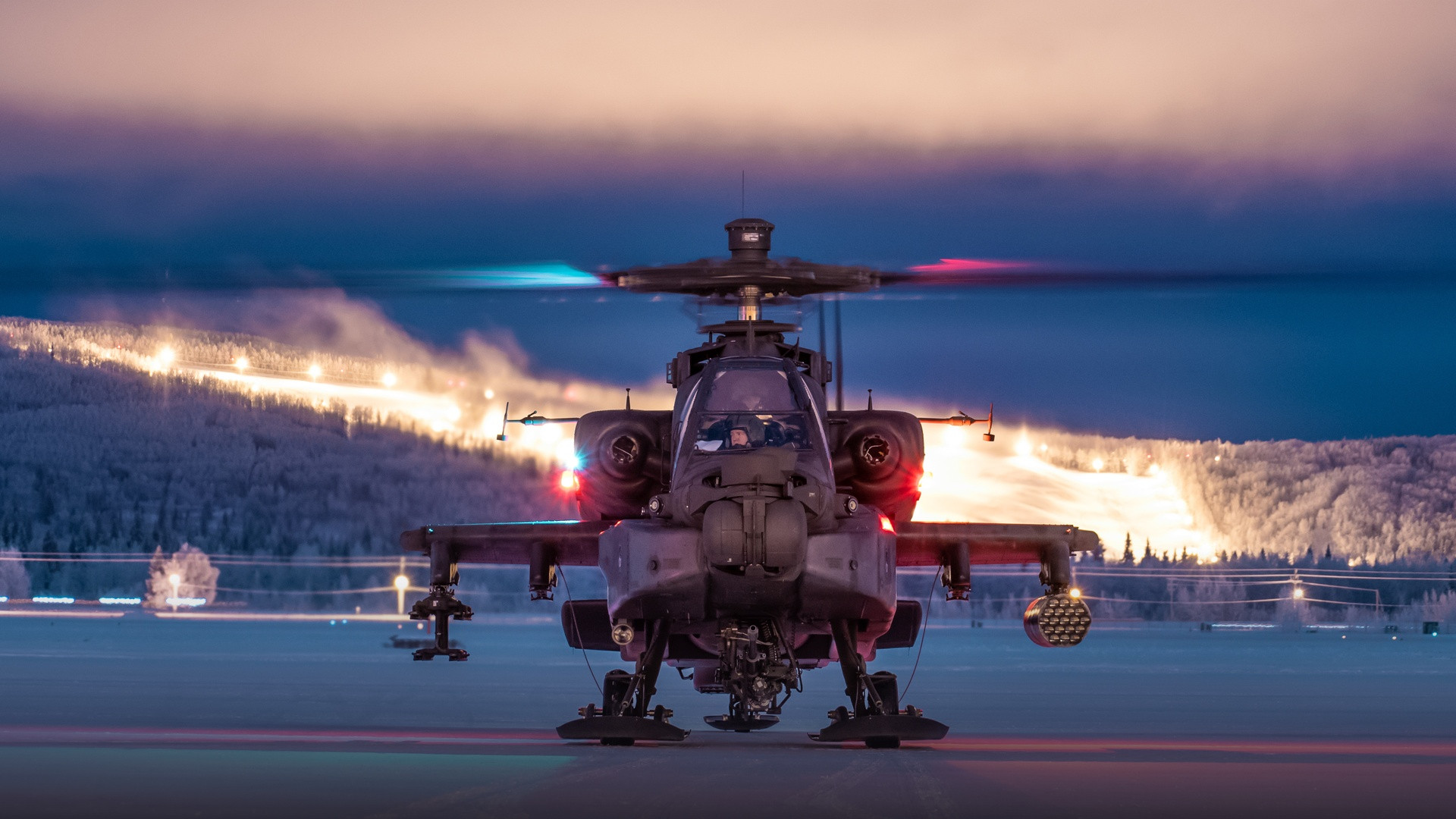 U.S. Army AH-64 Apache attack helicopter facing forward on a snow-covered airfield at twilight with cockpit illuminated.