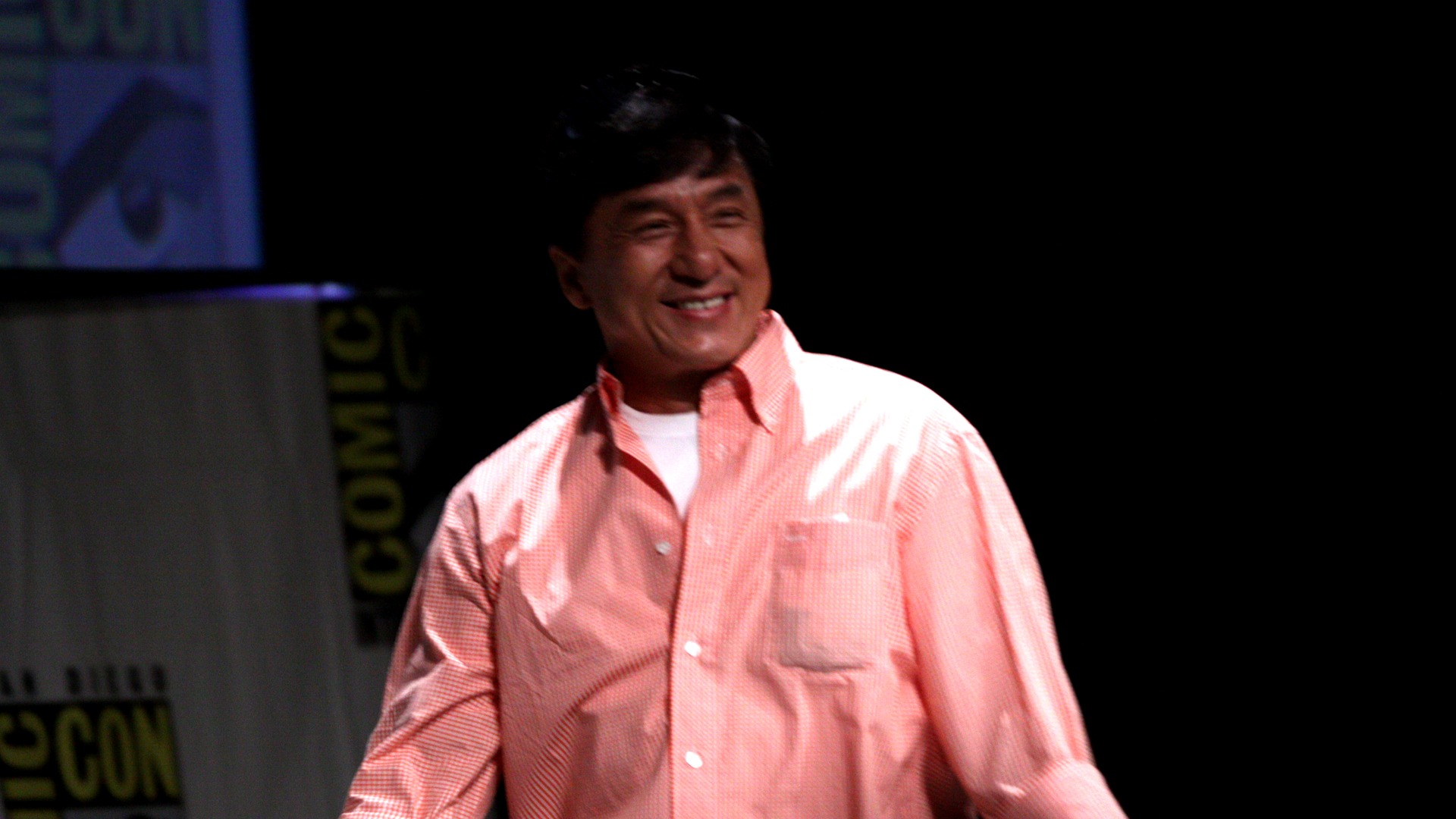 Martial arts icon Jackie Chan smiling and waving to fans at a red carpet event.