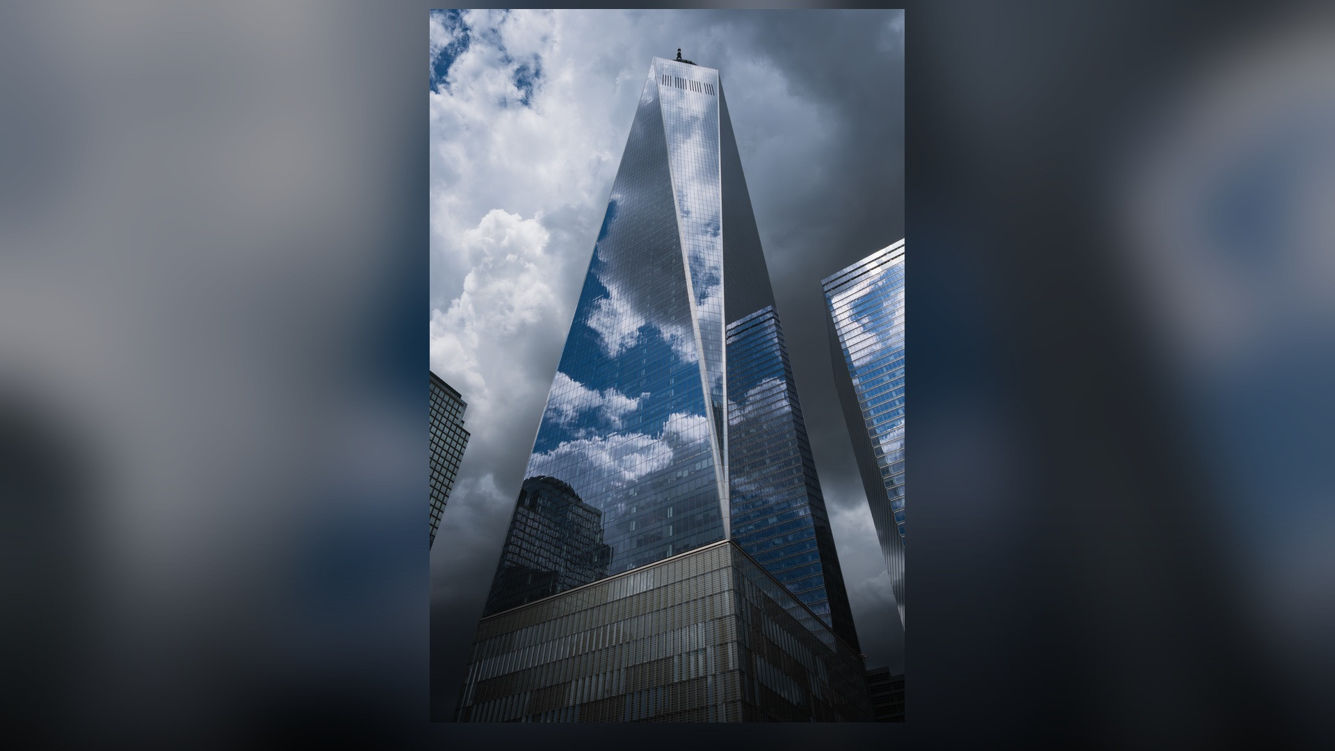 A vertical architectural photograph of One World Trade Center with its shining glass facade and spire tip.