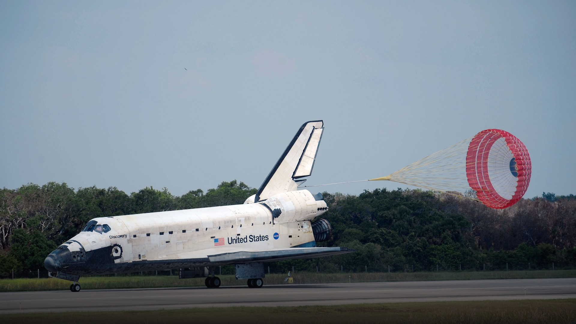 Space Shuttle Discovery touching down on a military airfield runway with red and white drag chute deployed.