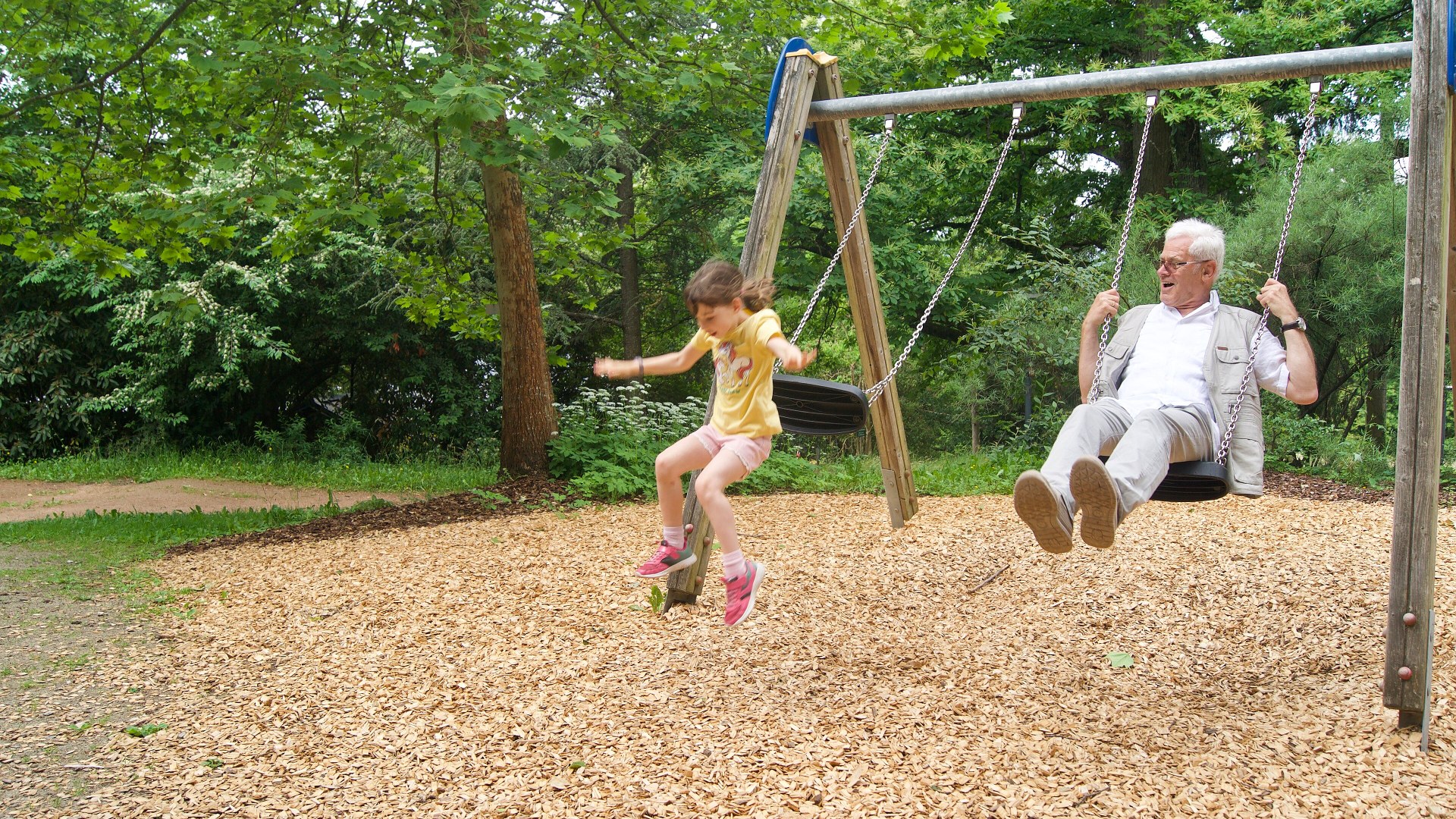 A grandfather and his granddaughter playing on outdoor swings together.