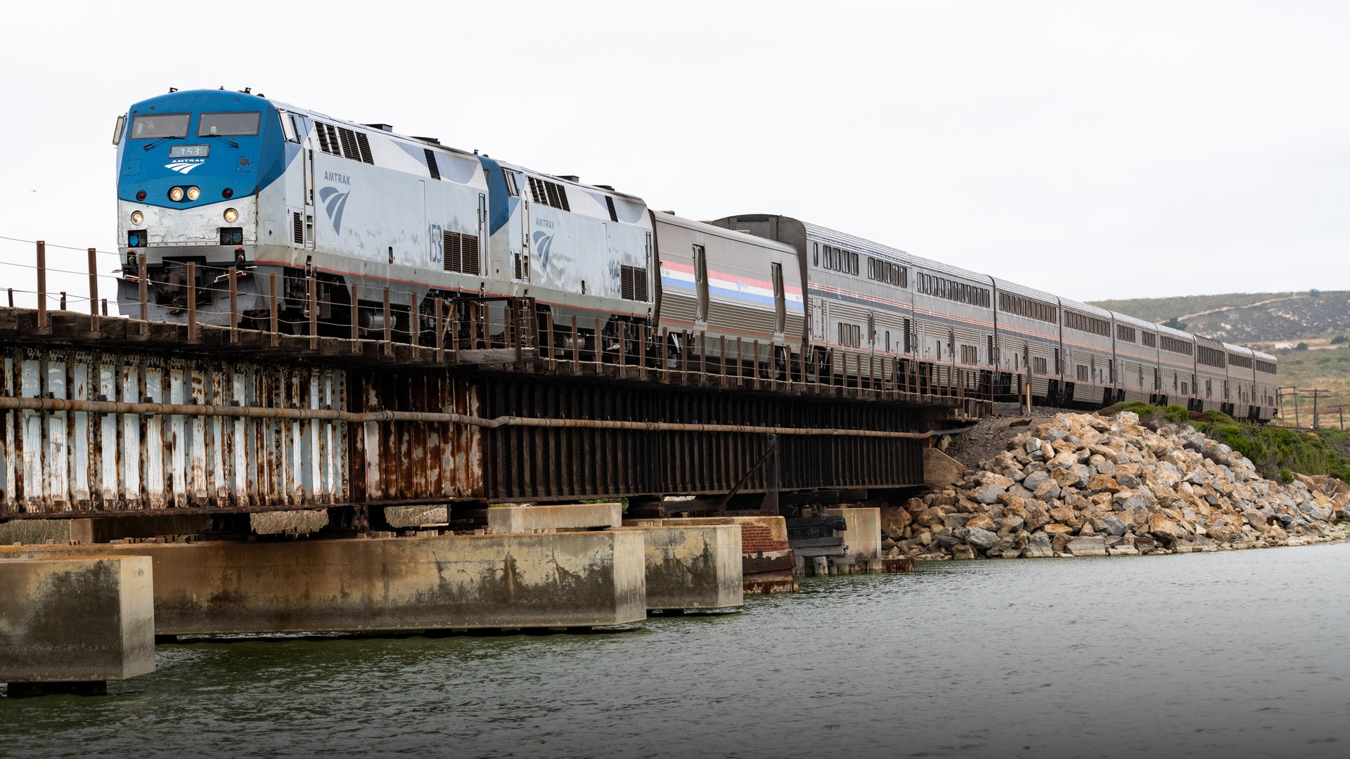 Silver double-decker Amtrak Coast Starlight train crossing a railway trestle bridge over tidal waters on the California coast