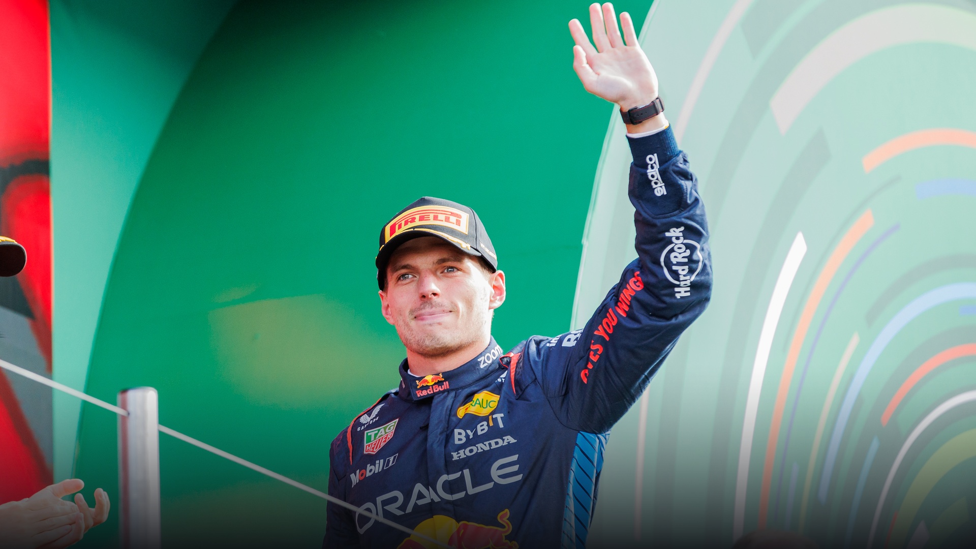 A joyous Max Verstappen waving triumphantly to cheering fans from the top of the podium.
