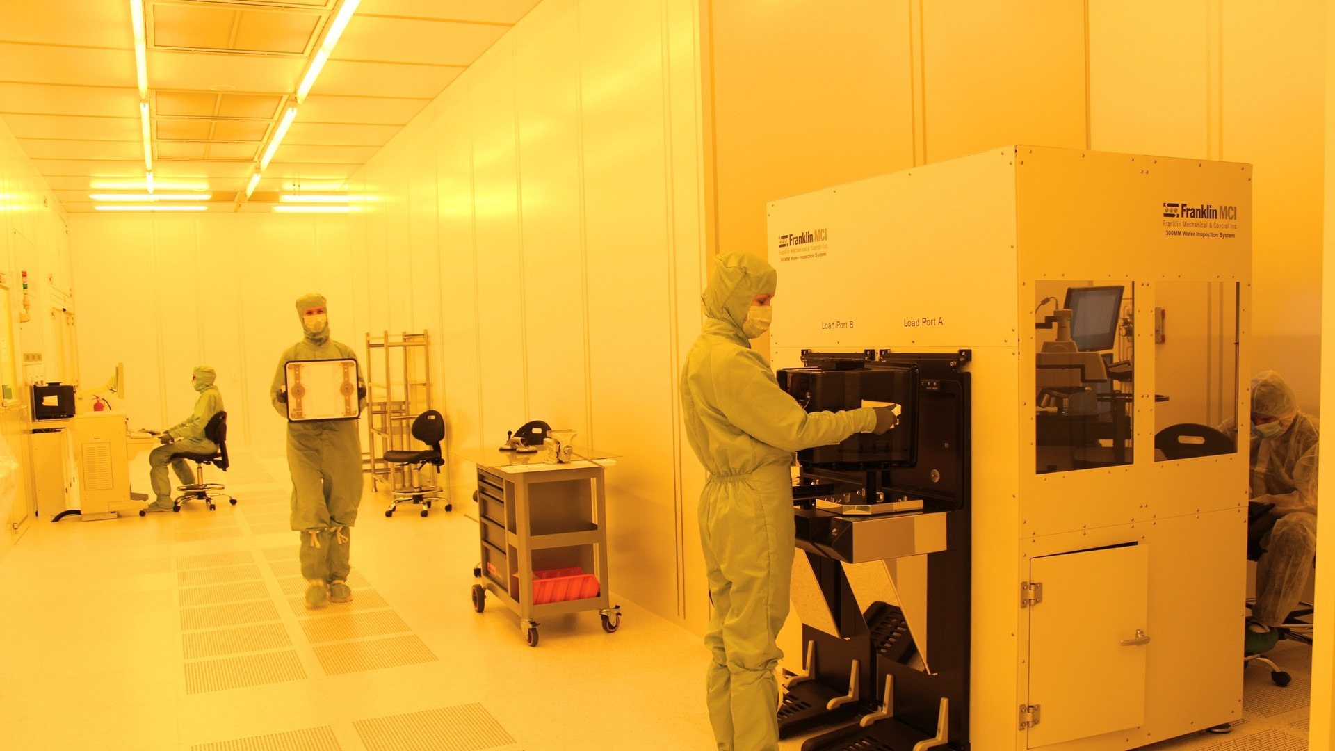 Cleanroom technicians in full white protective suits inspecting high-purity silicon wafers under ambient yellow lighting.