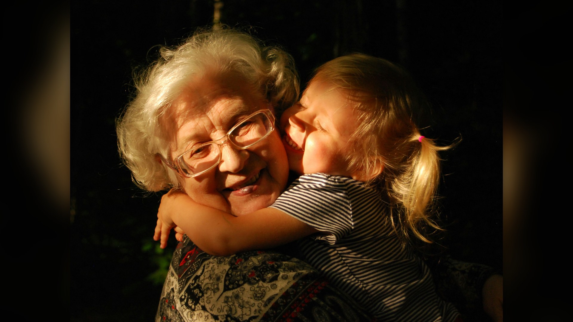 An elderly grandmother with glasses warmly hugging a young child.