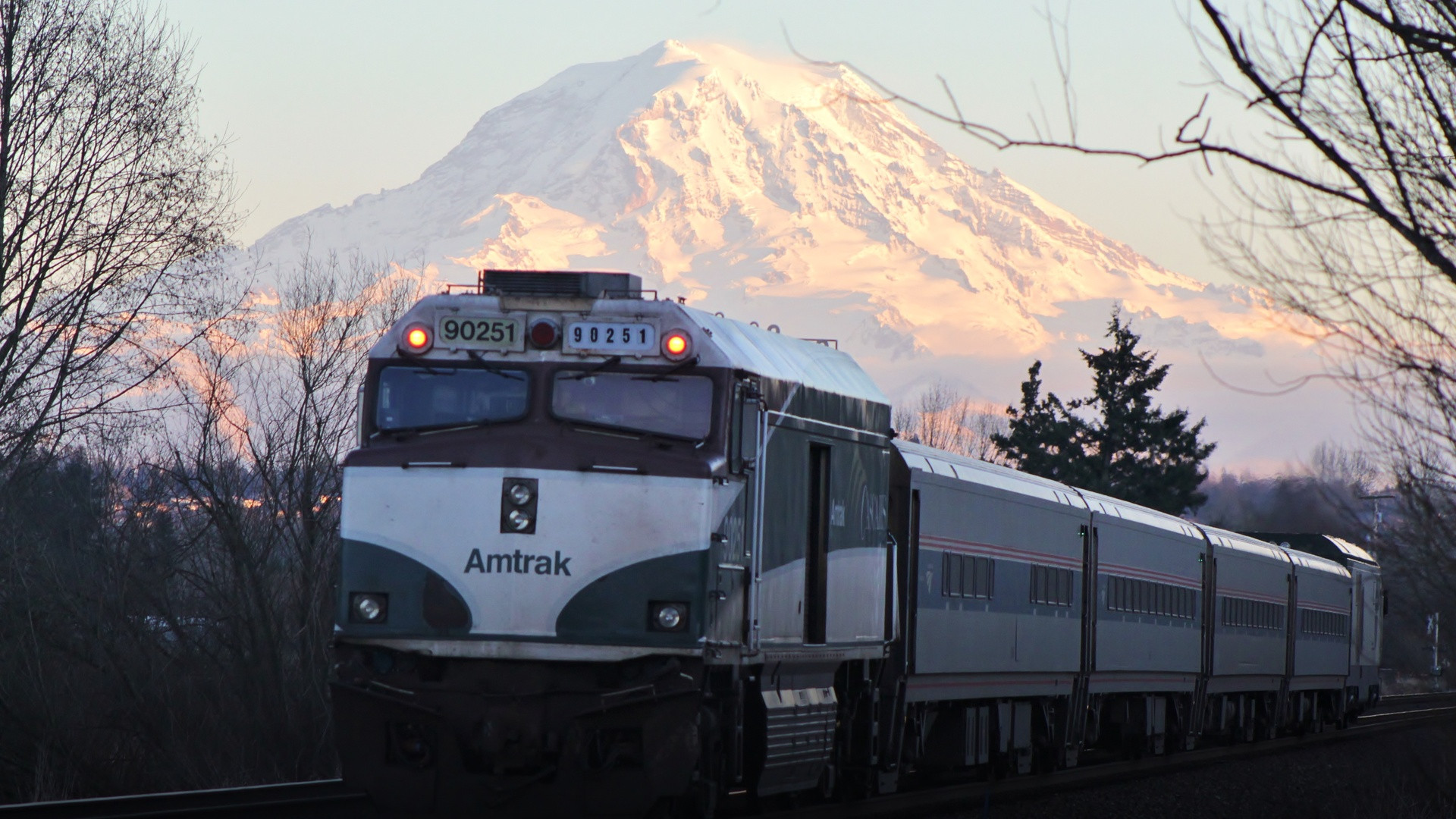 Amtrak Cascades passenger train traveling along tracks with the towering snow-covered summit of Mount Rainier in the background