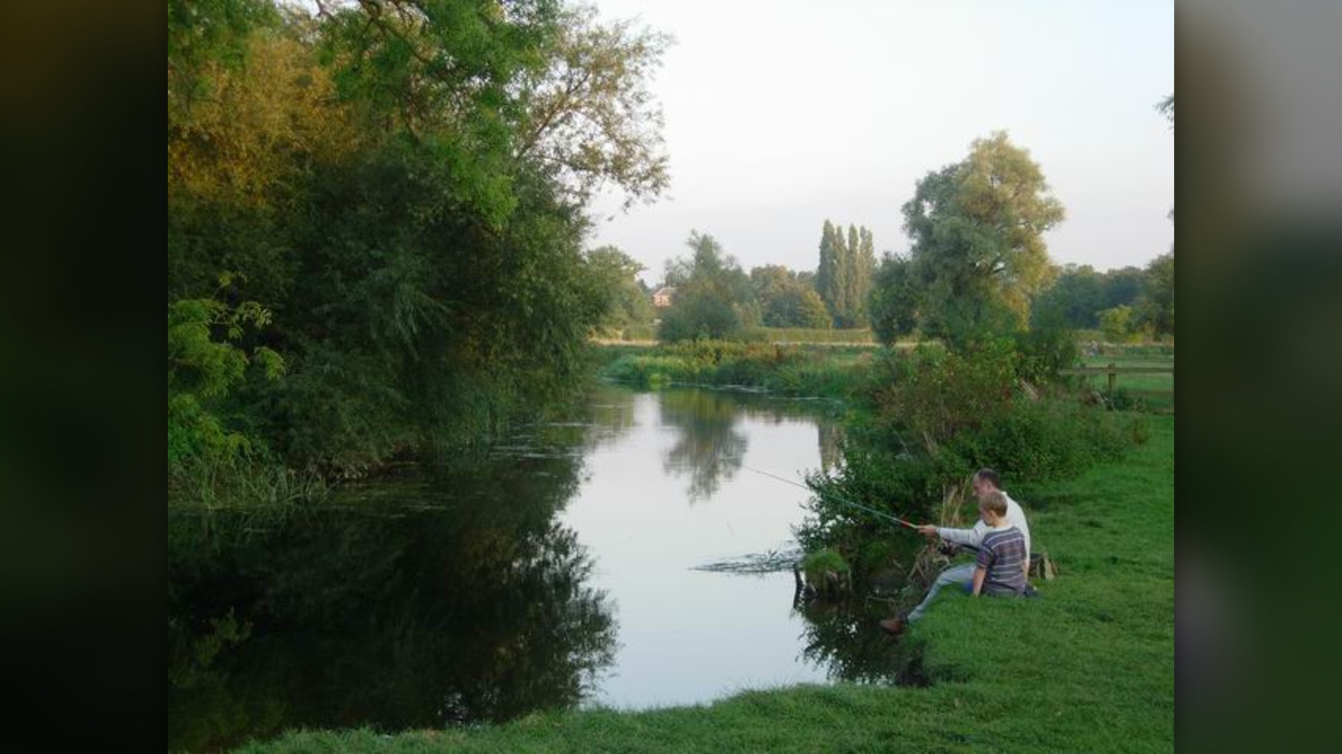 A grandfather and child fishing together along the riverbank in the evening.