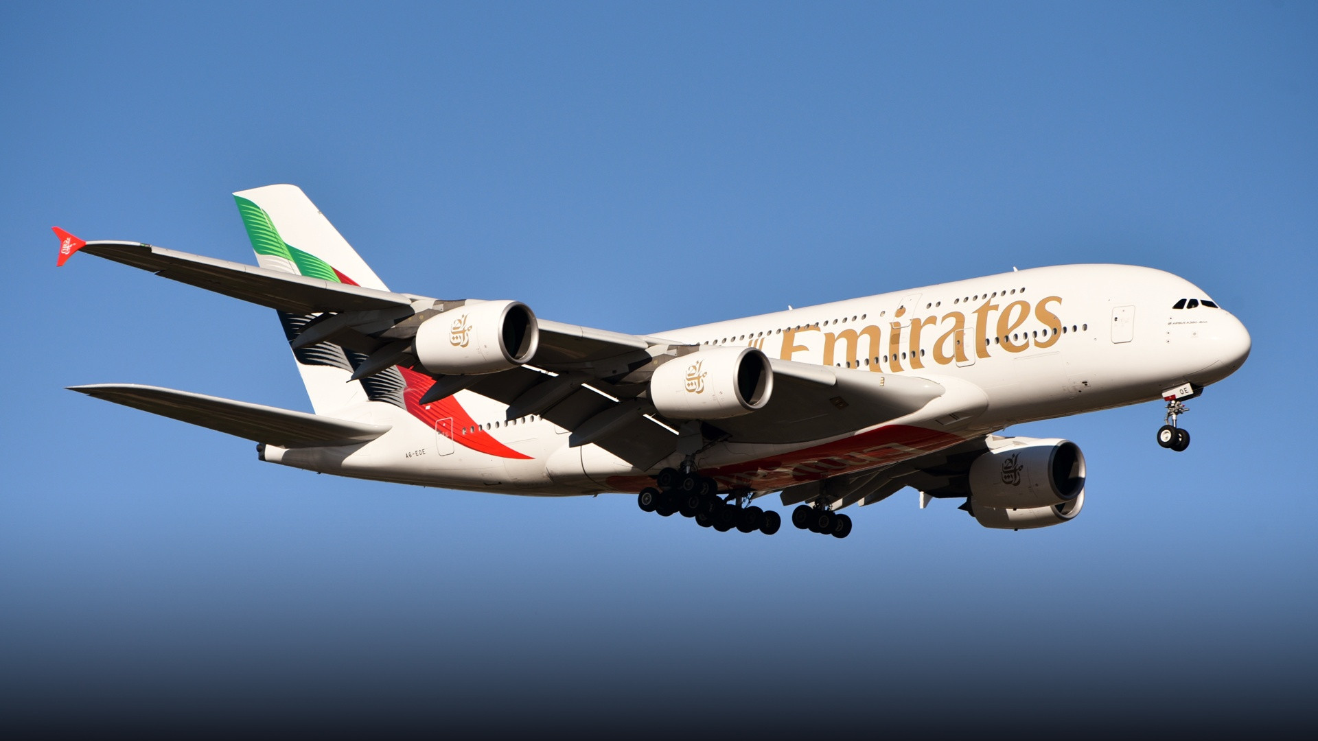 An Emirates Airbus A380-800 on final landing approach with gear deployed under a clear sky.
