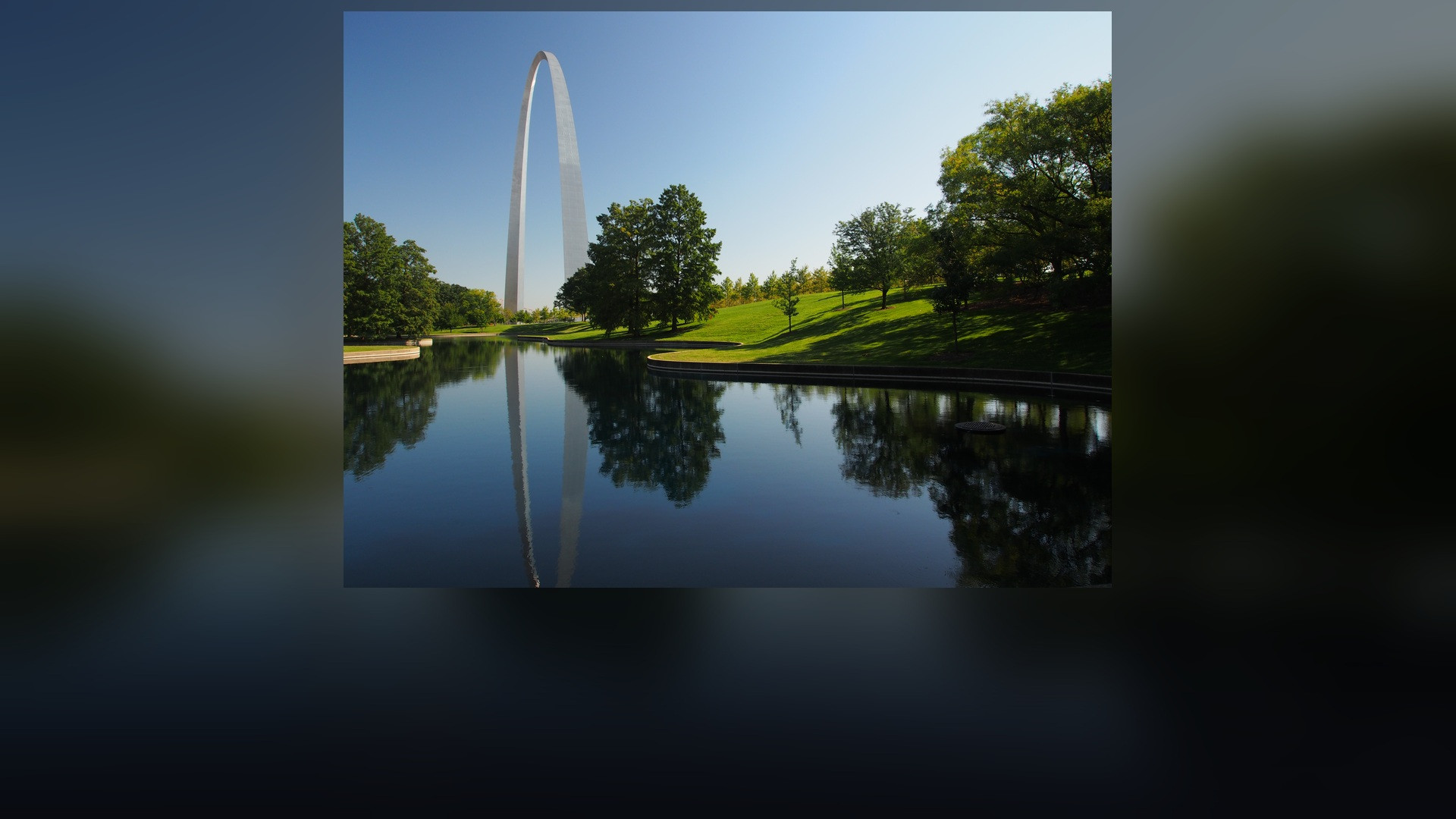 Exterior view of the towering stainless-steel Gateway Arch reflecting in the park's south reflection pond with green lawns and blue sky.