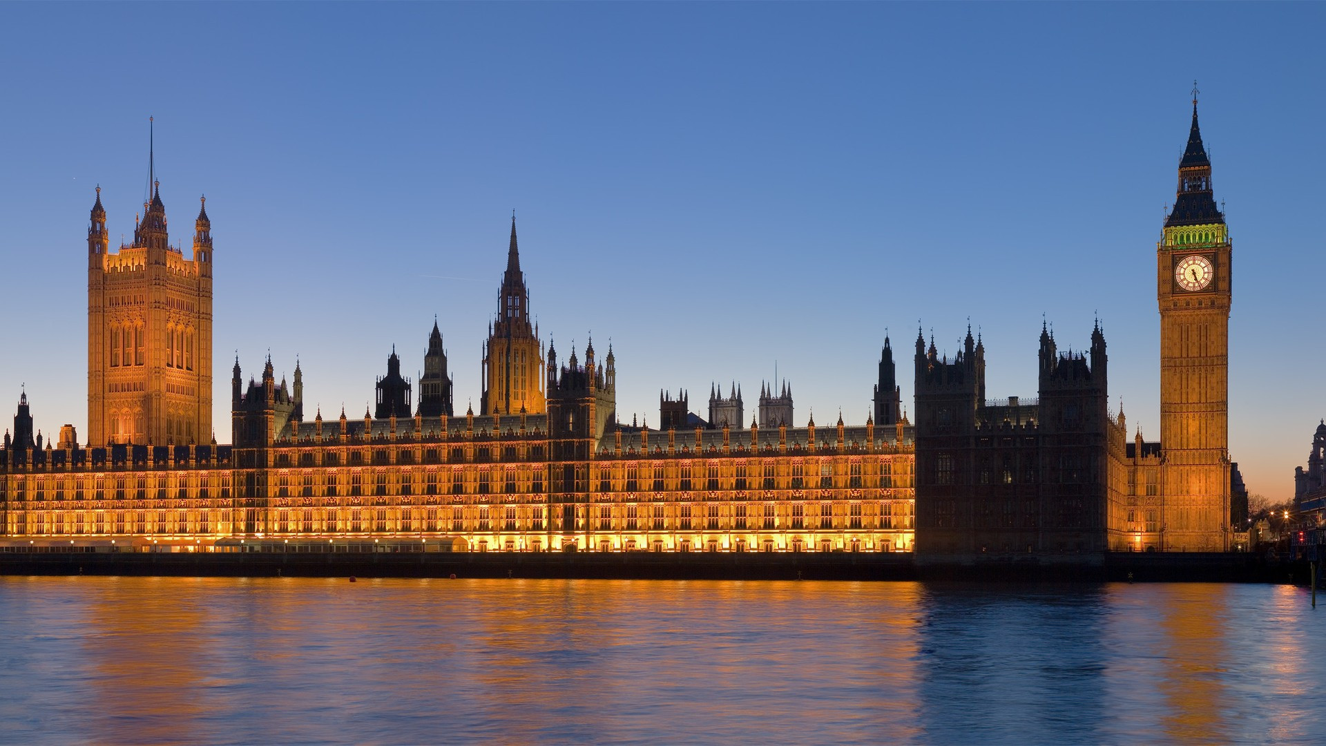The illuminated Gothic facade of the Palace of Westminster and Big Ben clock tower reflected across the River Thames at dusk.