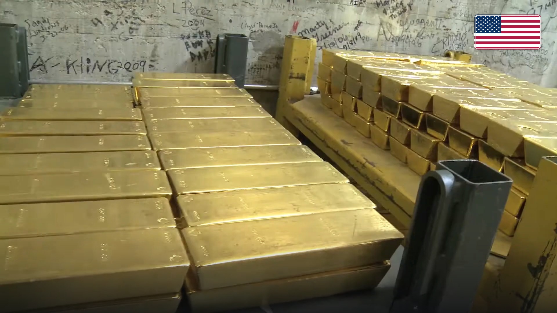 Pallets and stacks of gold bullion bars inside the United States Mint Bullion Depository at West Point. Official national flag badge in top right.