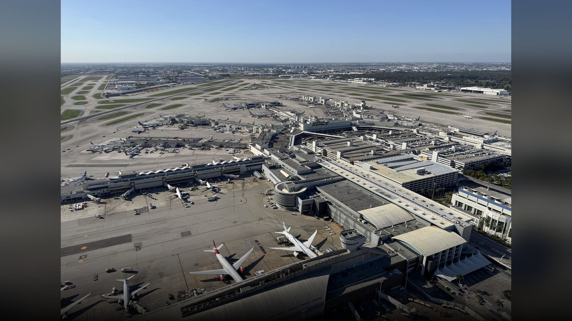 Widebody passenger airliners parked at the modern concourse gates of Miami International Airport under sunny skies.