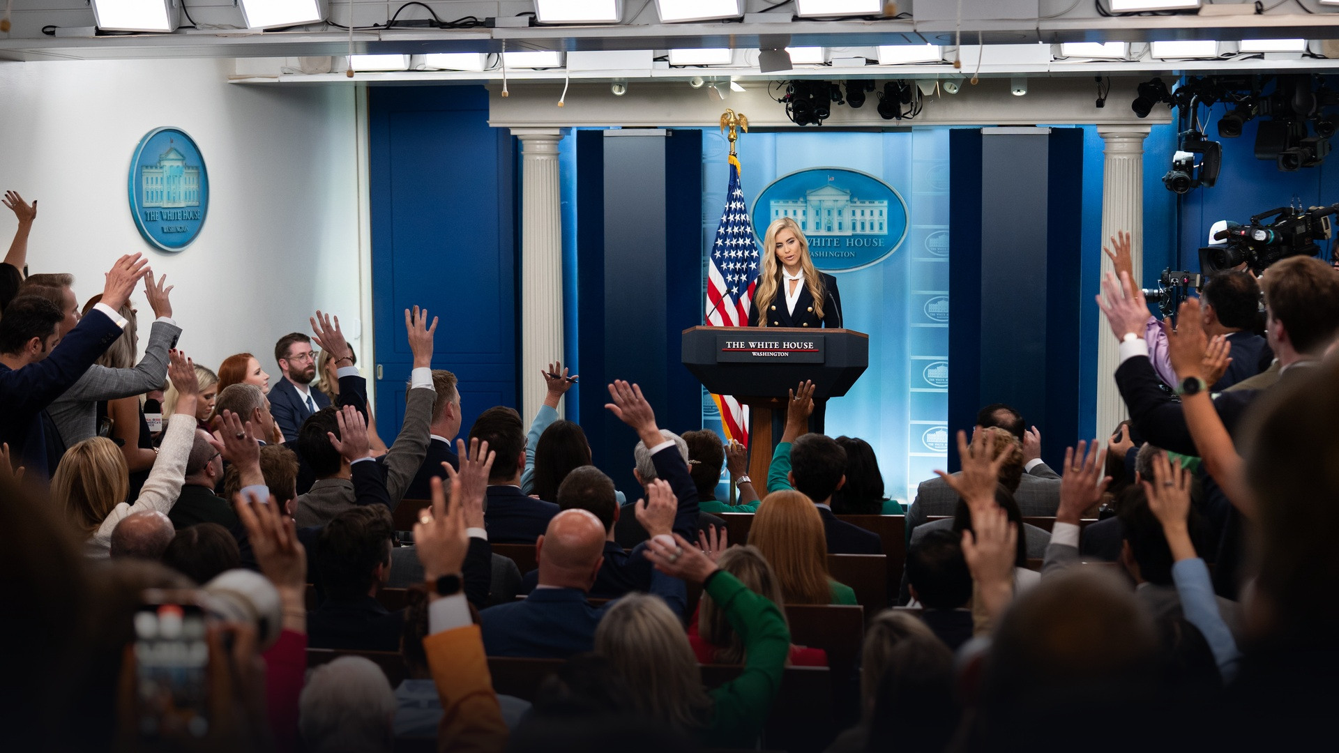 White House Press Secretary Katie Zacharia at the podium in the James S. Brady Press Briefing Room facing raised hands from reporters on October 13, 2026.
