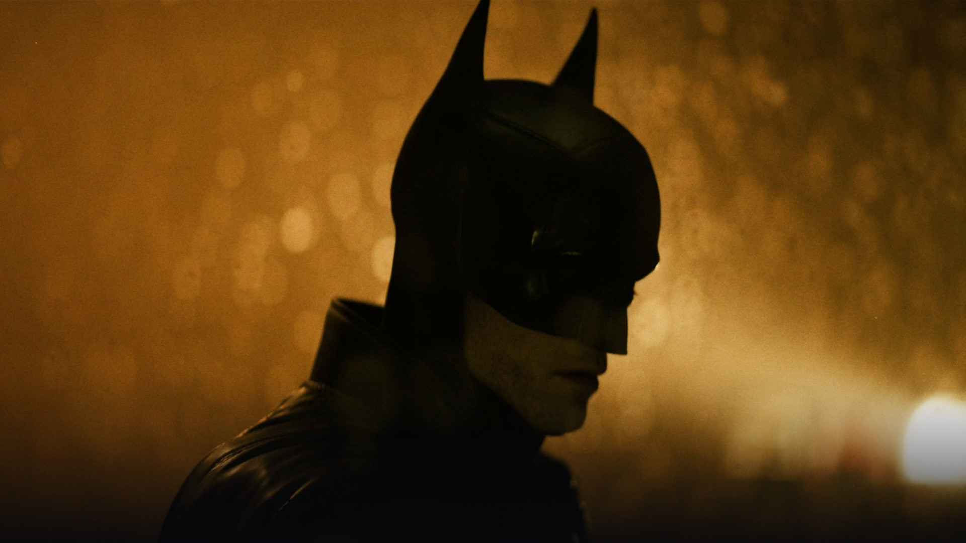 Robert Pattinson in the rugged Batsuit standing amid falling snow in dark Gotham City.