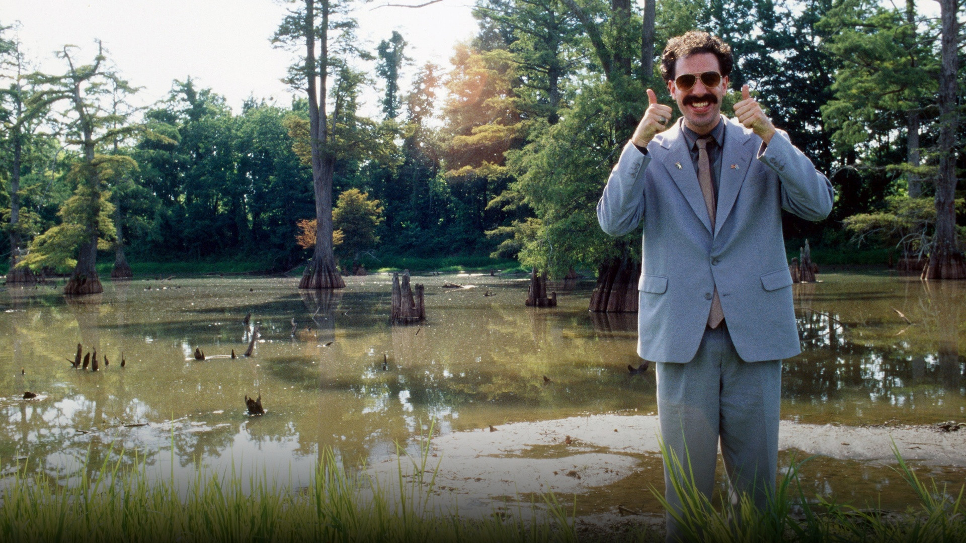 Sacha Baron Cohen as Kazakh television reporter Borat Sagdiyev giving an energetic double thumbs-up in his gray suit.