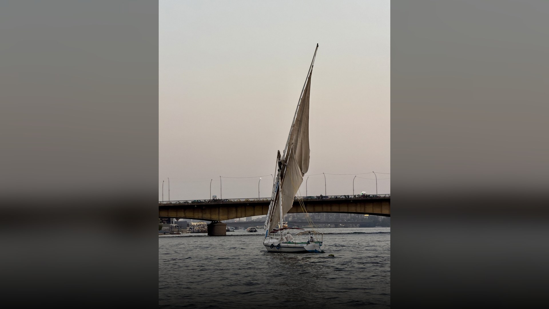 Traditional wooden felucca sailboat gliding across the calm Nile River with Cairo city buildings in the background.