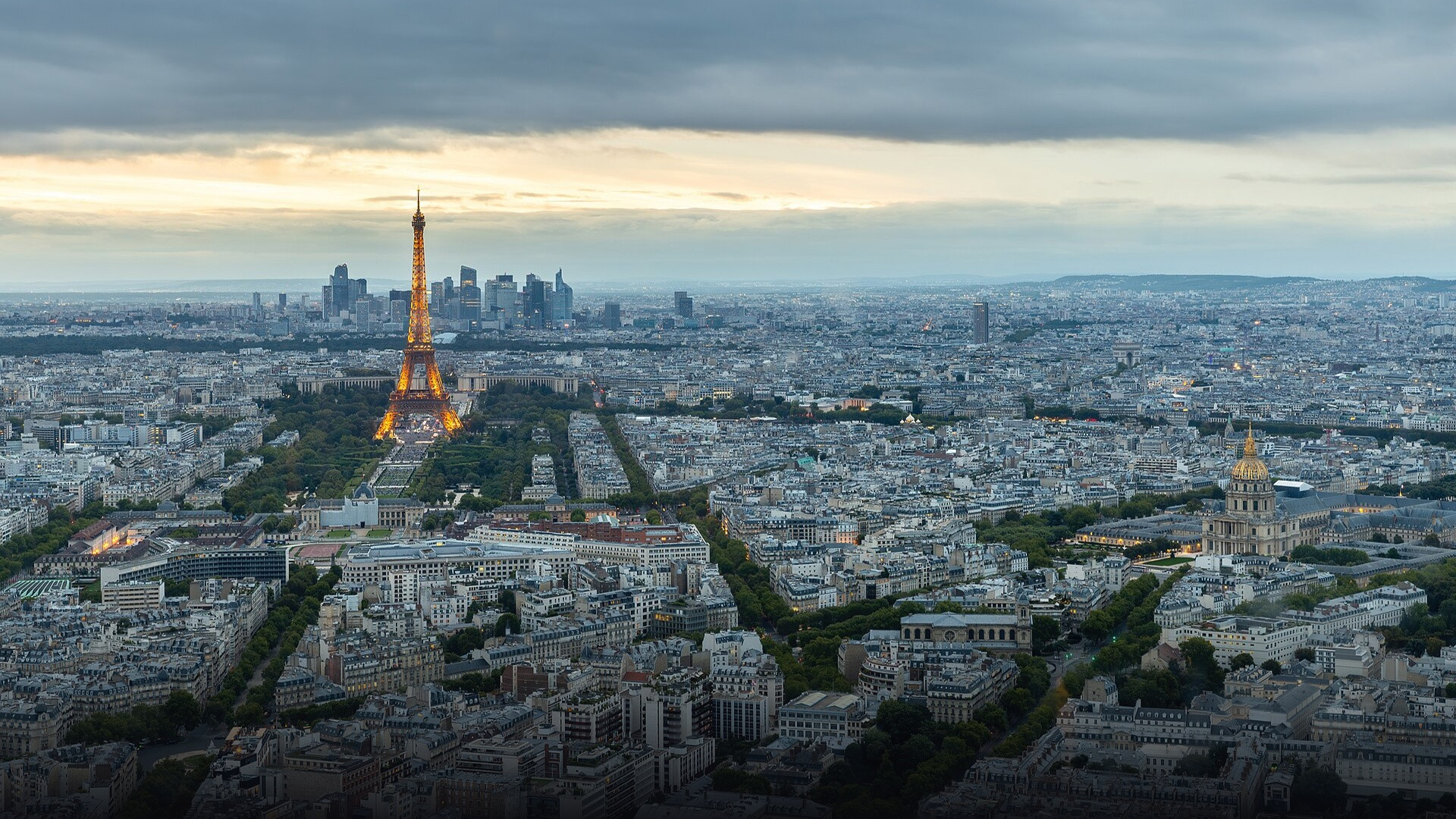 Panoramic twilight view over the Paris skyline featuring the illuminated Eiffel Tower.