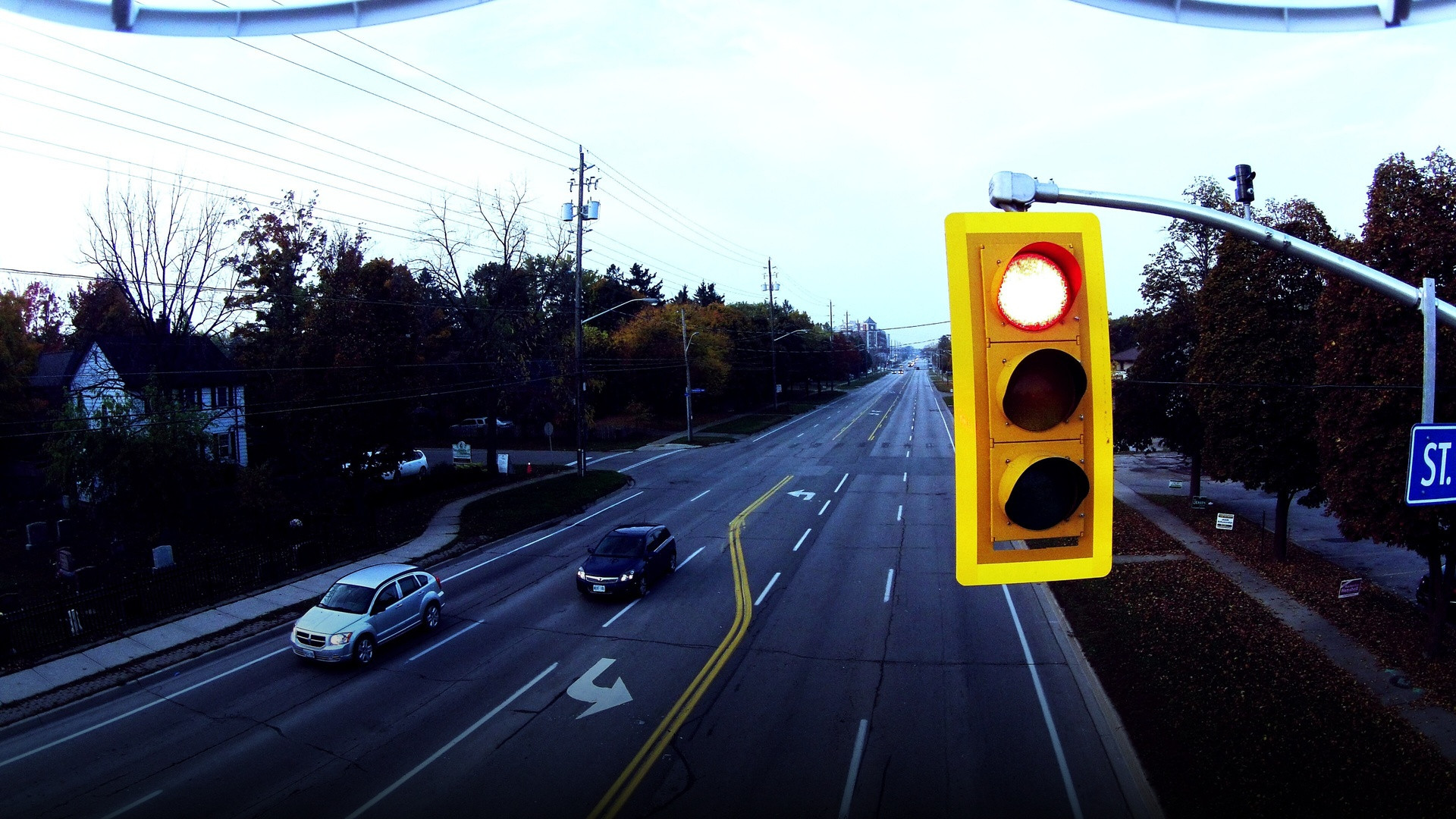 An elevated view of an American roadway intersection showing a prominent overhead yellow traffic signal glowing red above traffic lanes.