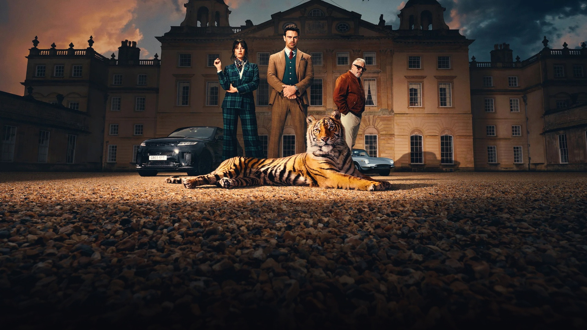 Theo James and Kaya Scodelario standing stylishly before an English country manor with a Bengal tiger resting in foreground.