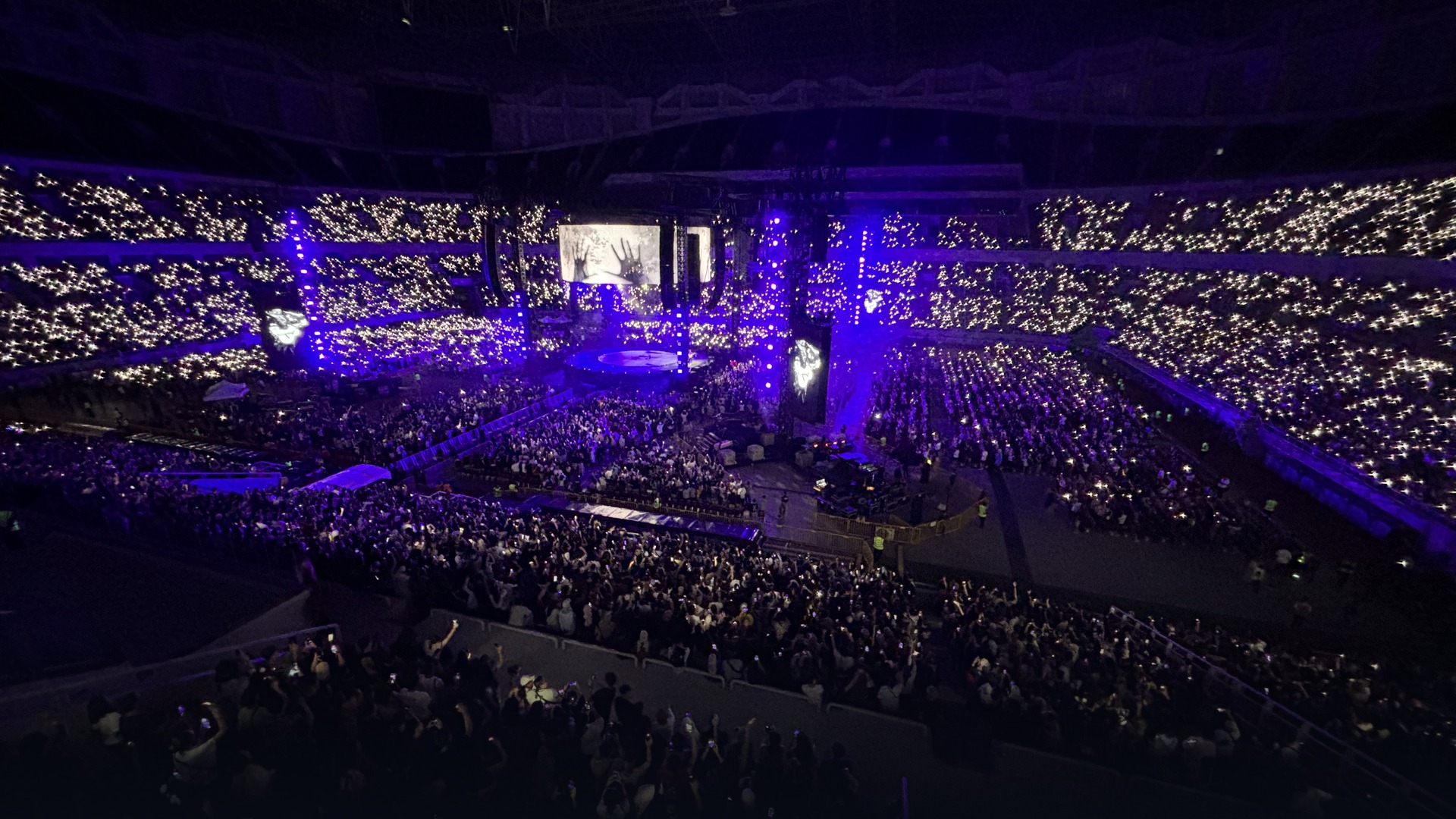 Vast stadium concert crowd illuminated under massive 360-degree stage structures.