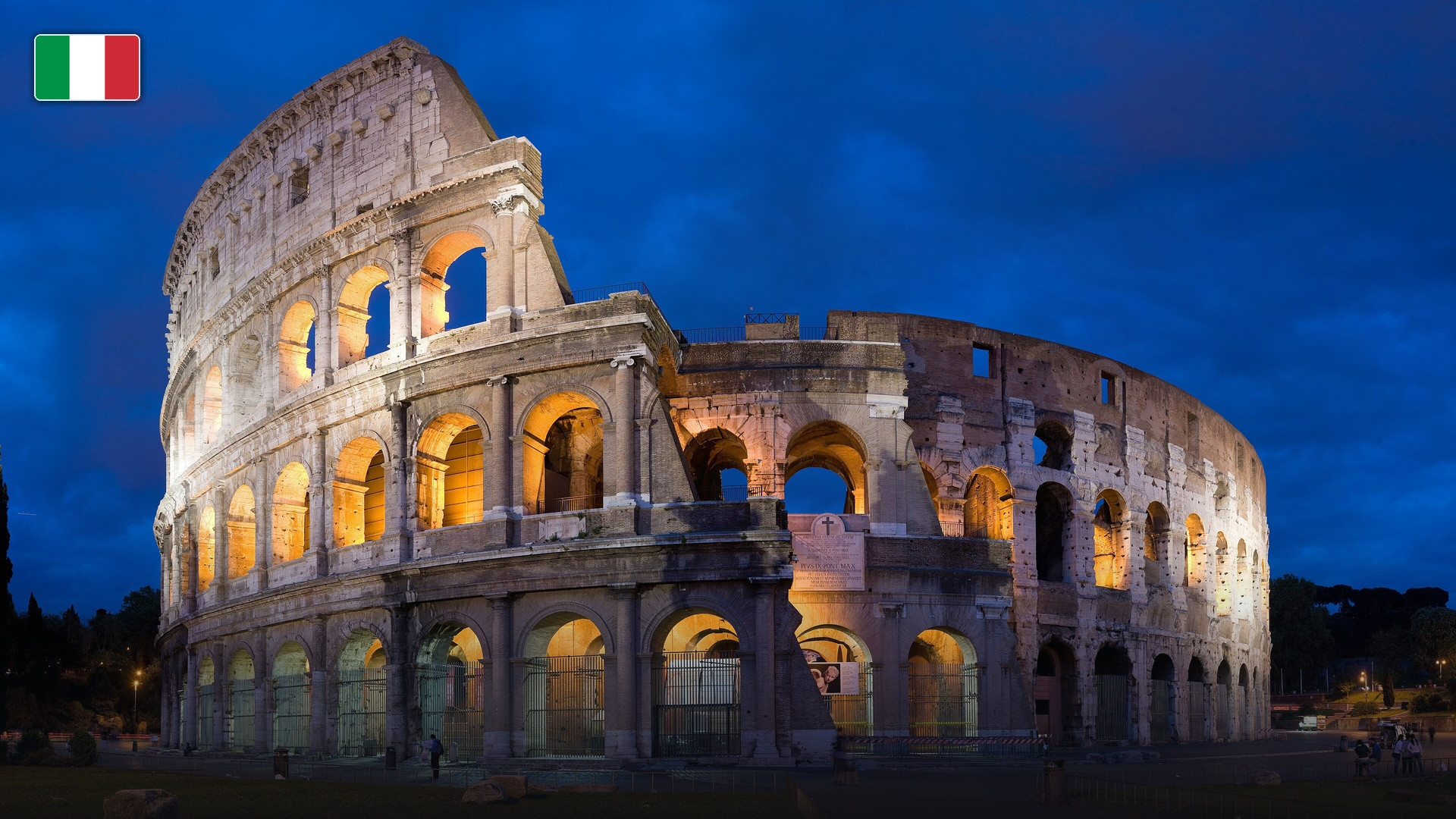 The ancient Roman Colosseum illuminated under clear skies, with the flag of Italy.