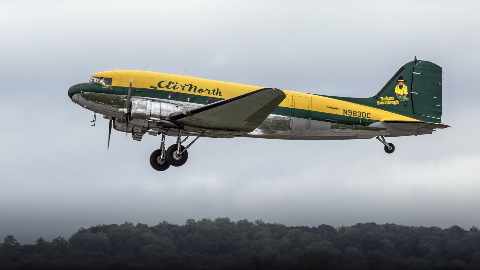 An Air North Douglas DC-3 in yellow and green livery flying low on landing approach against cloudy hills.