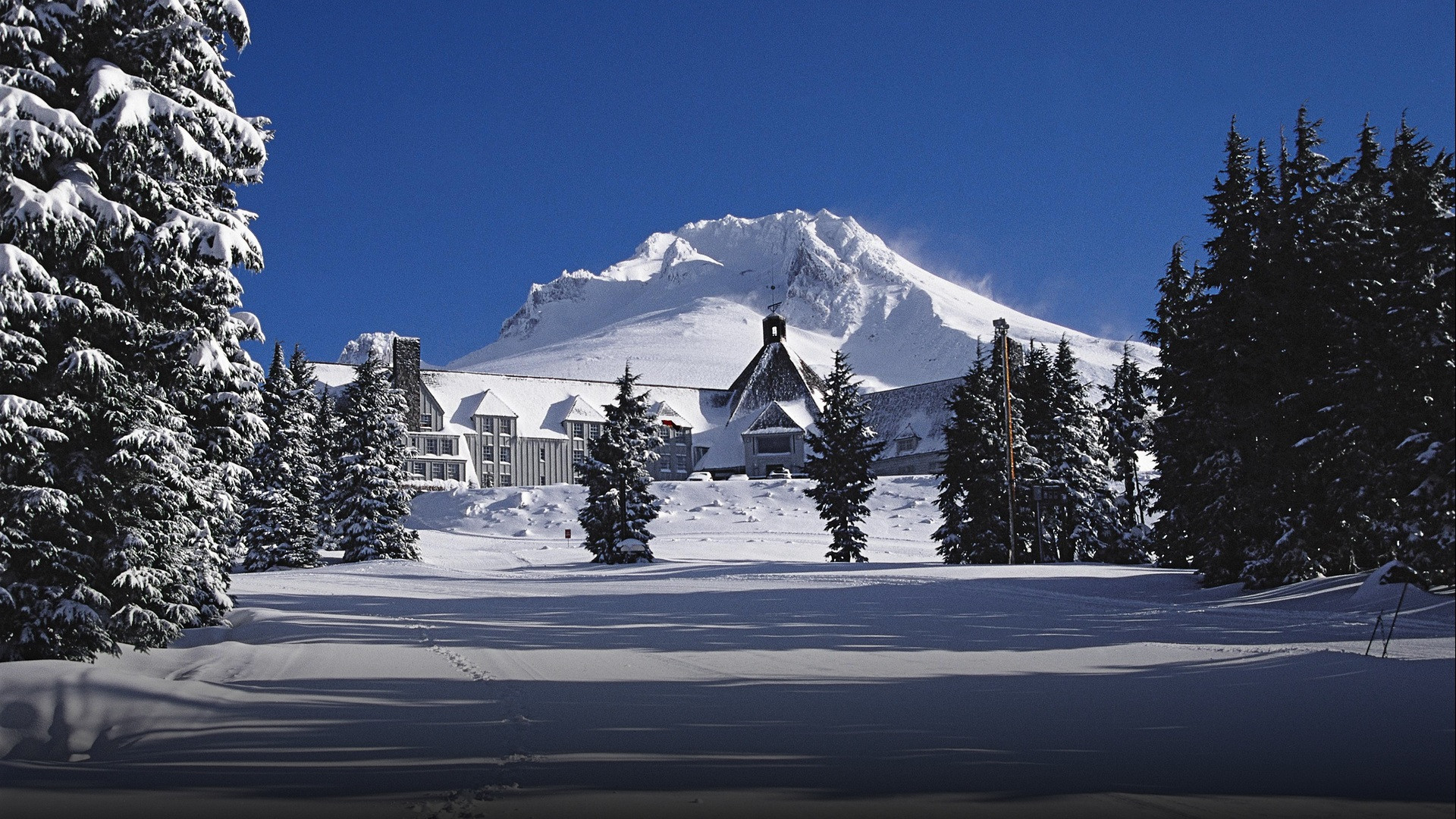 Timberline Lodge blanketed in deep winter snow with the snow-capped peak of Mount Hood rising under a brilliant blue sky.