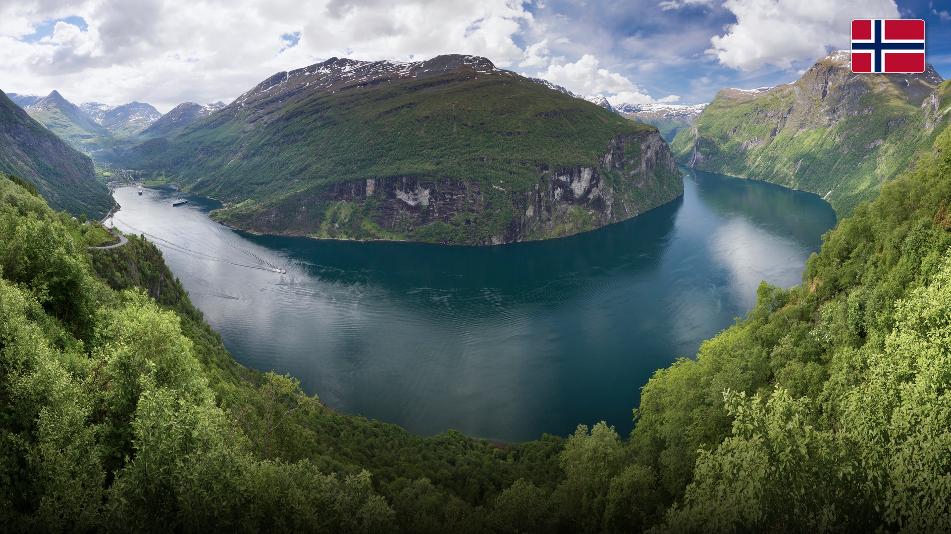 Panoramic view over Geirangerfjord surrounded by sheer green cliffs and cascading waterfalls.