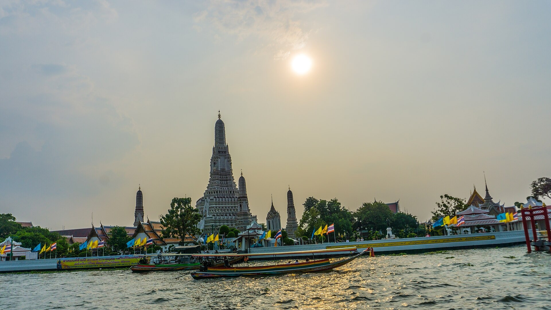 Wat Arun Temple of Dawn with its iconic central prang tower and longtail boats on the Chao Phraya River at sunset.