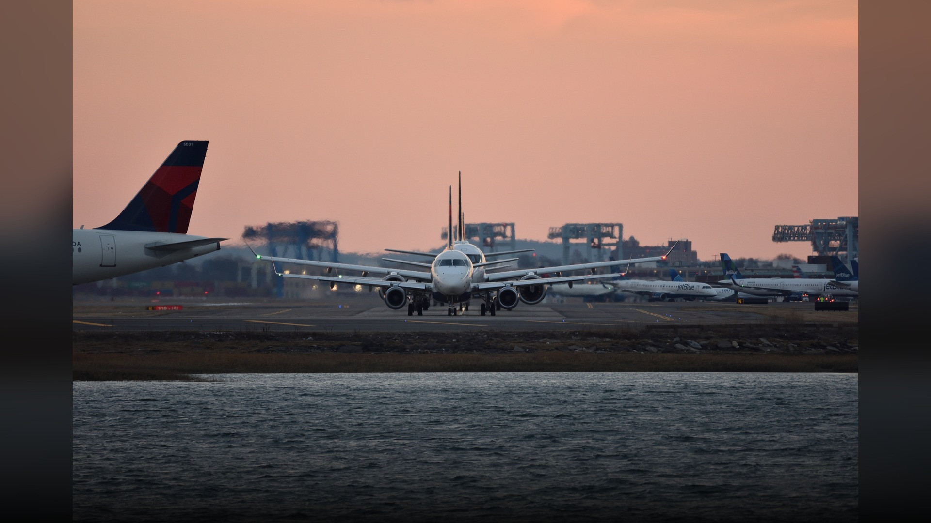 Multiple commercial aircraft lining up along the taxiway at Boston Logan International Airport with harbor waters in background.