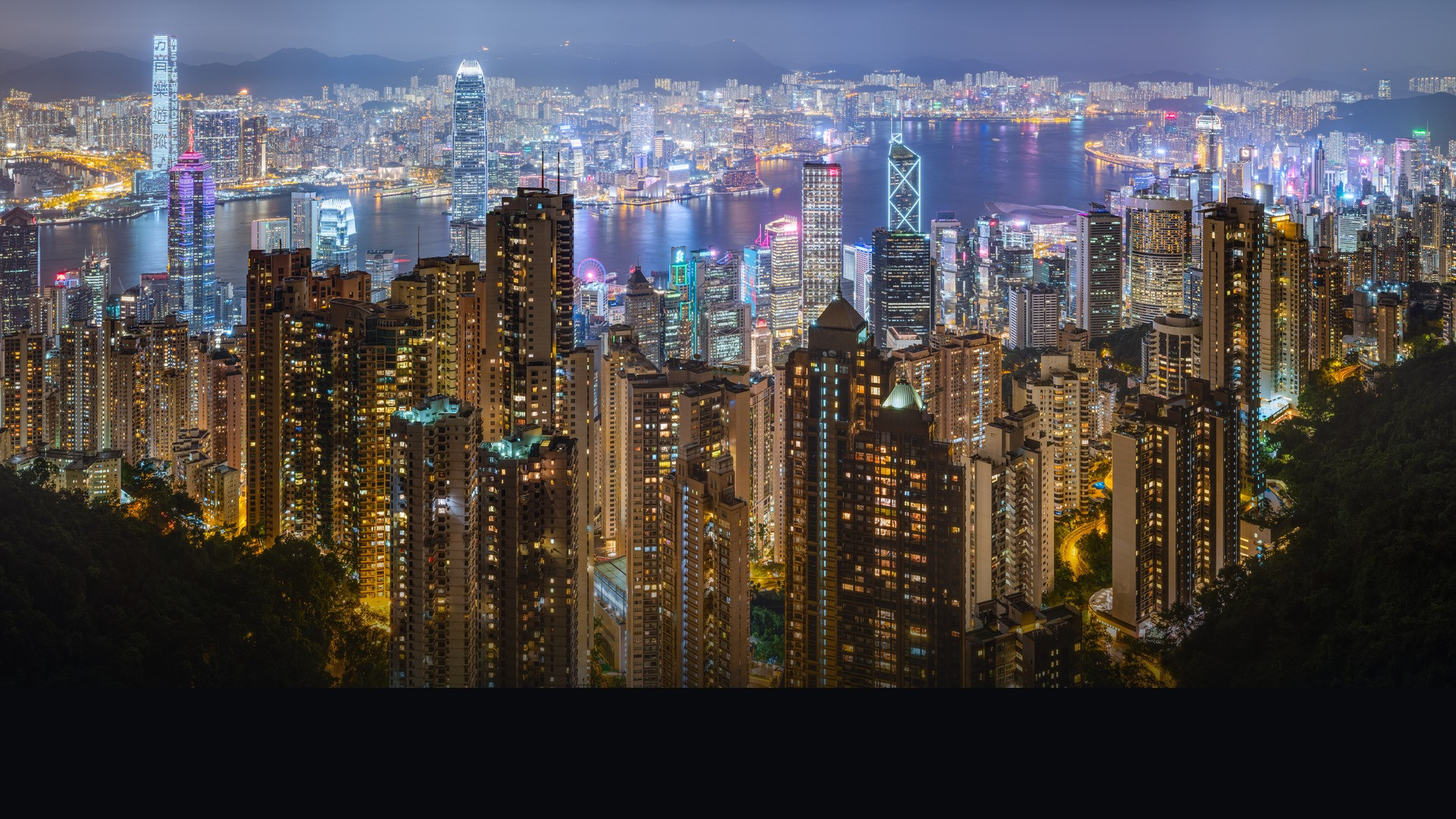 Panoramic evening view of the illuminated Hong Kong Island skyline across Victoria Harbour.