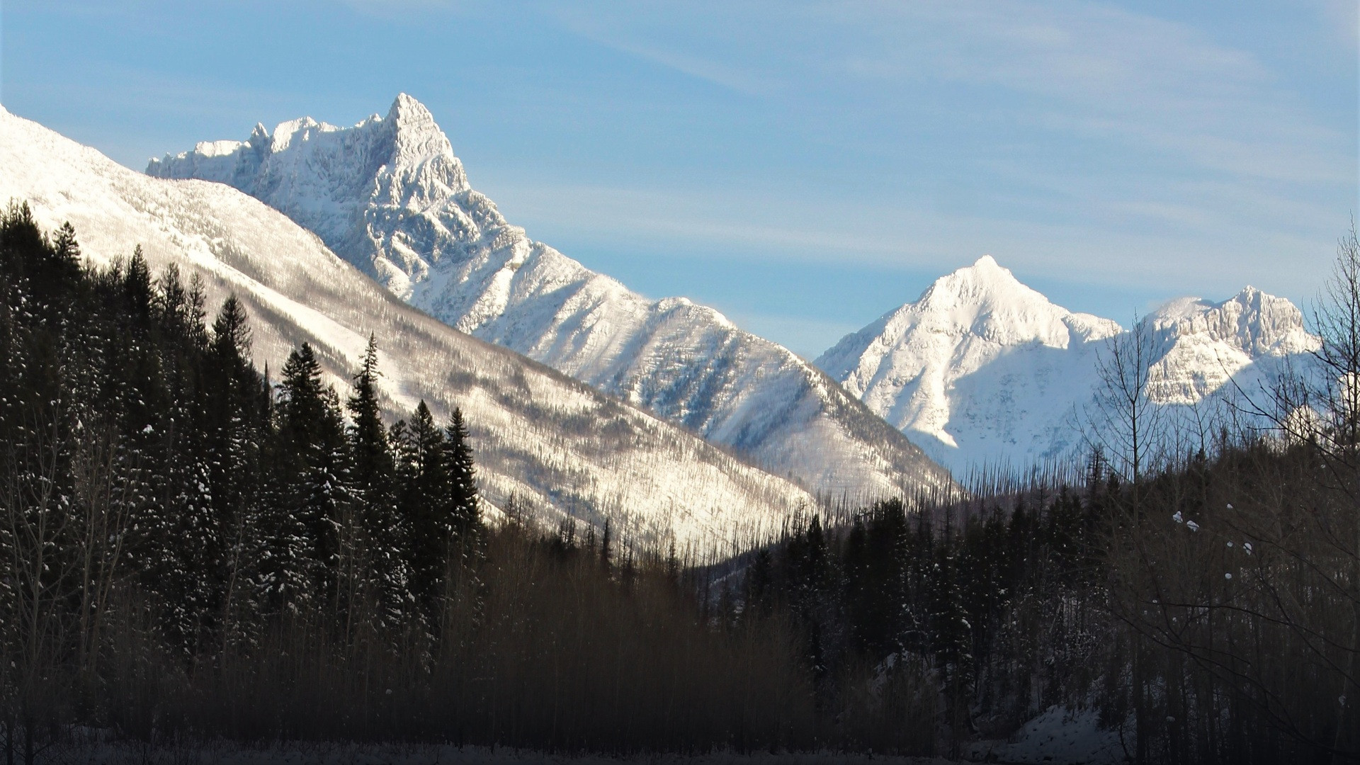 Jagged mountain peaks covered in snow overlooking a tranquil, frosty lake in Glacier National Park.