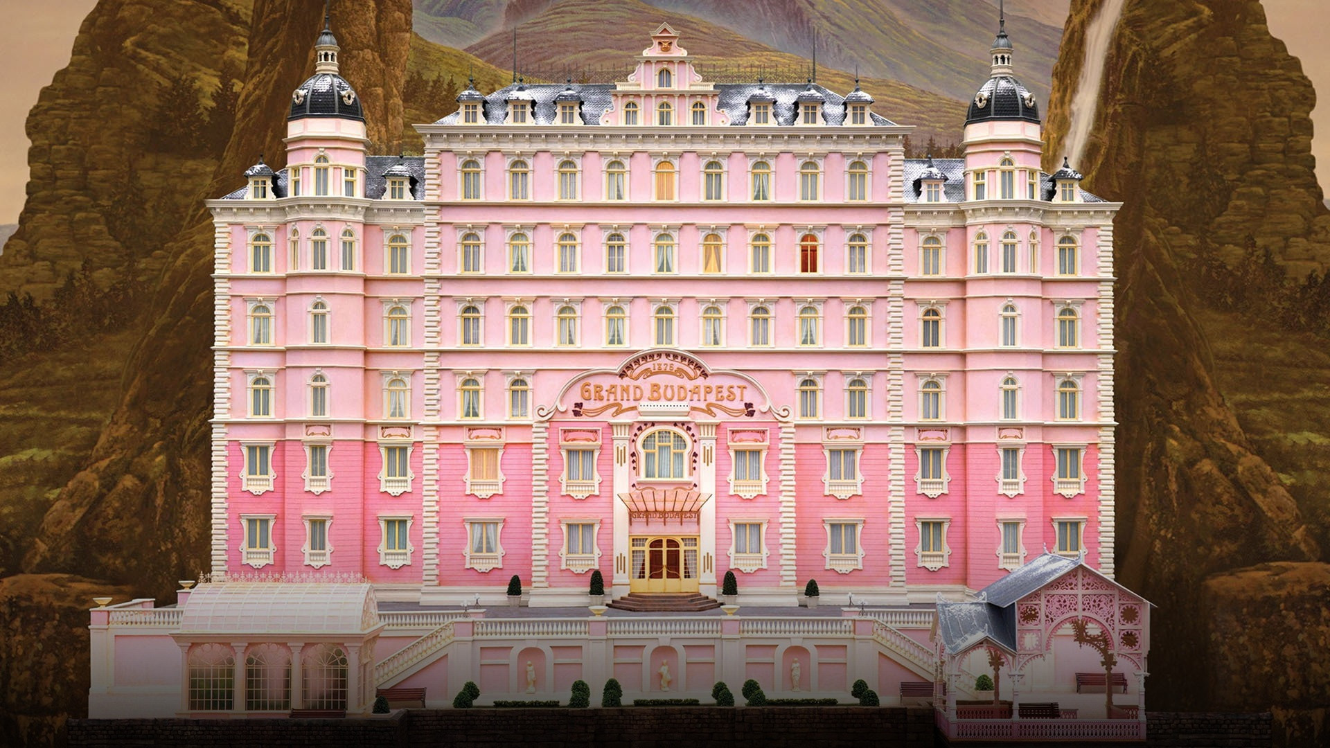 The symmetrical pastel-pink facade of The Grand Budapest Hotel against misty alpine cliffs.