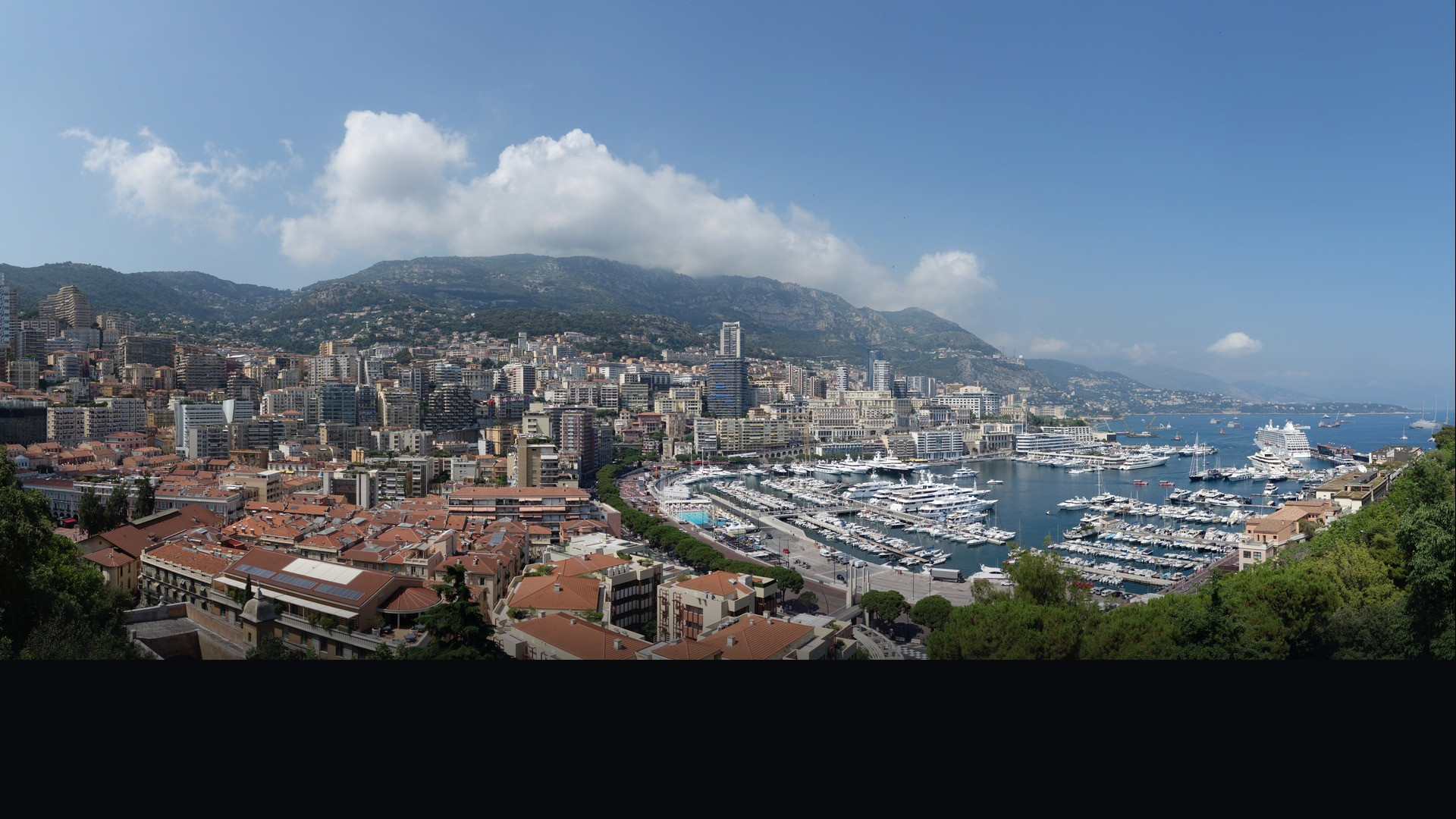 Sun-drenched panoramic view of Monaco's Port Hercule with luxury yachts and coastal cliffs.