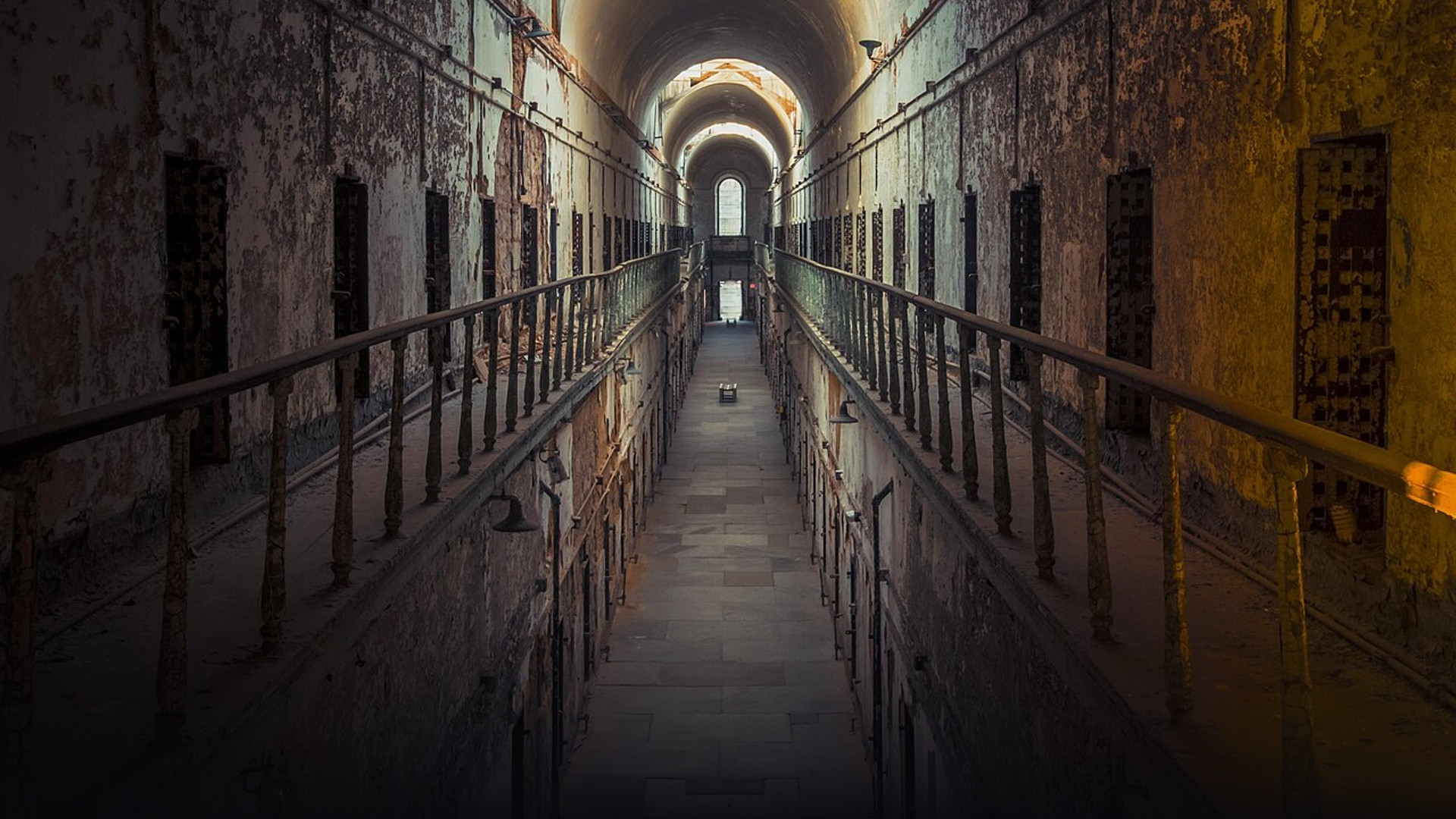 The towering two-story radial cellblock corridor of Eastern State Penitentiary featuring rows of iron cell doors, decaying plaster, and vaulted skylights.
