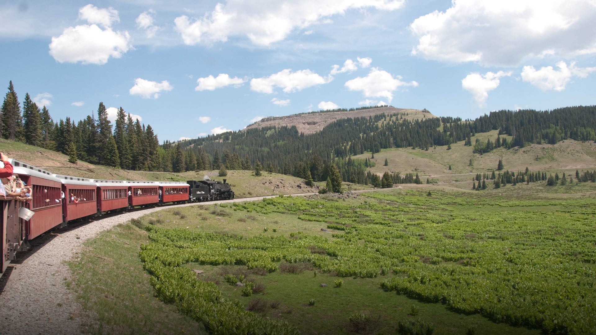 Vintage narrow-gauge steam train with red passenger coaches winding through a high-altitude green mountain valley