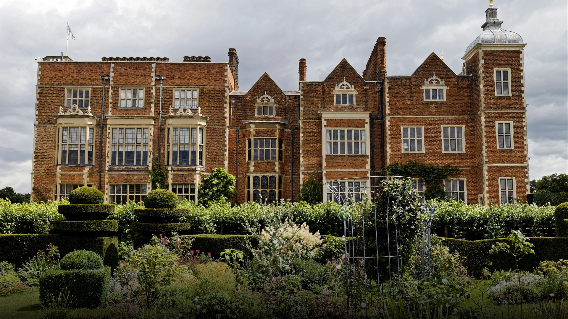 The red-brick Jacobean wing and carved stone gables of Hatfield House viewed past manicured topiary hedges.