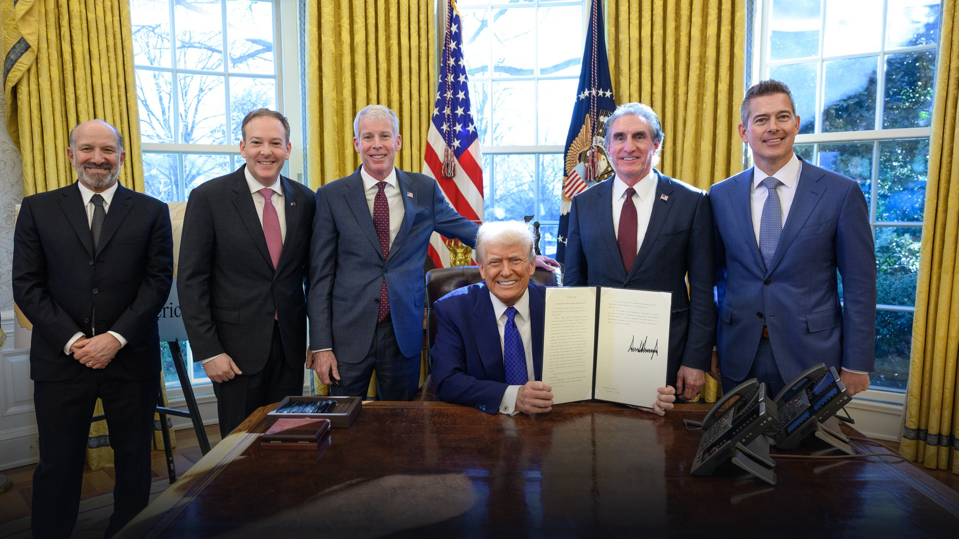 President Donald Trump seated at the Resolute Desk in the Oval Office surrounded by senior administration officials.