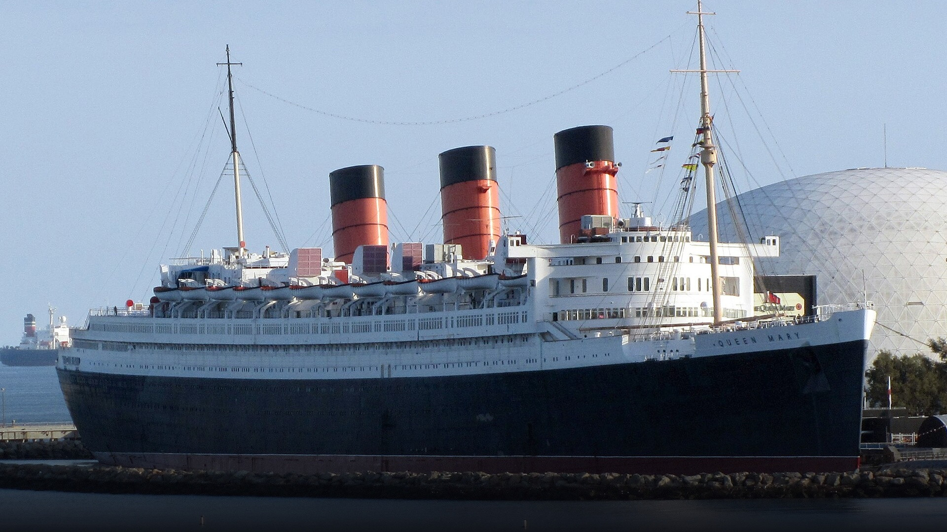 Portside profile of the iconic vintage ocean liner RMS Queen Mary docked in Long Beach harbor with its massive red-and-black funnels.