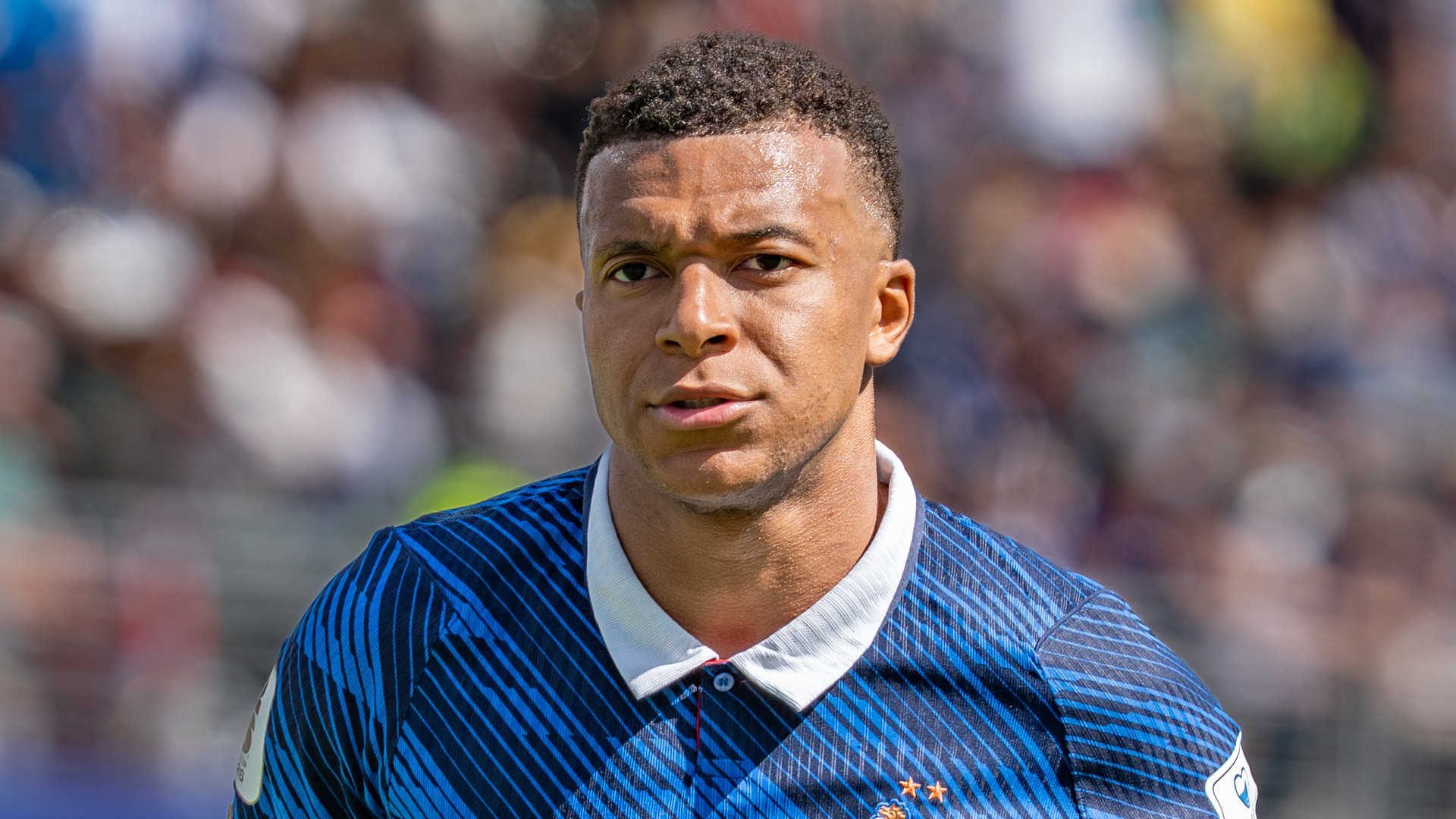 Close-up photograph of Kylian Mbappé in France's blue kit looking forward with concentration under stadium lights.