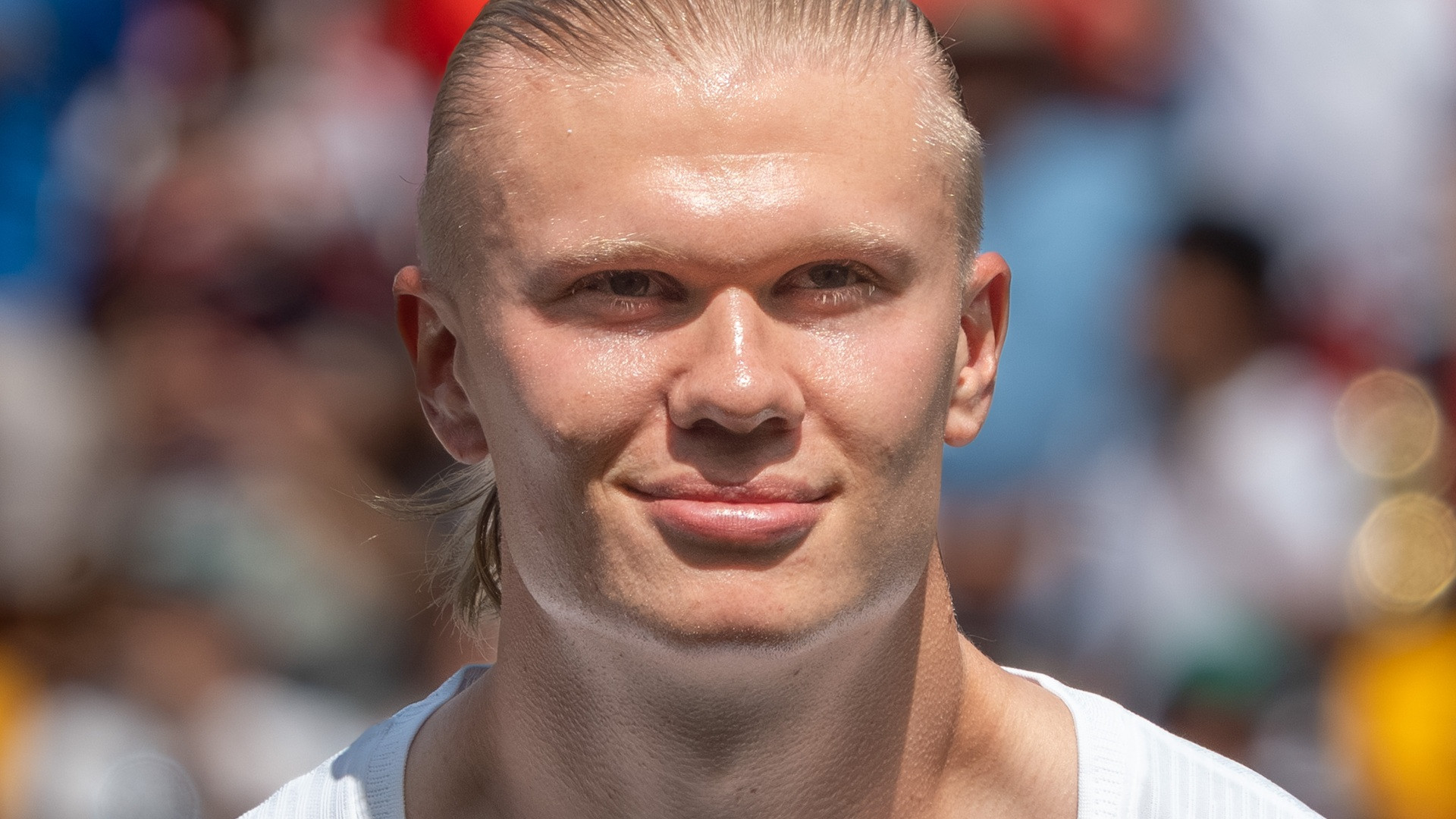 Close-up photograph of Erling Haaland smiling slightly with his hair tied back, framed against the stadium stands.