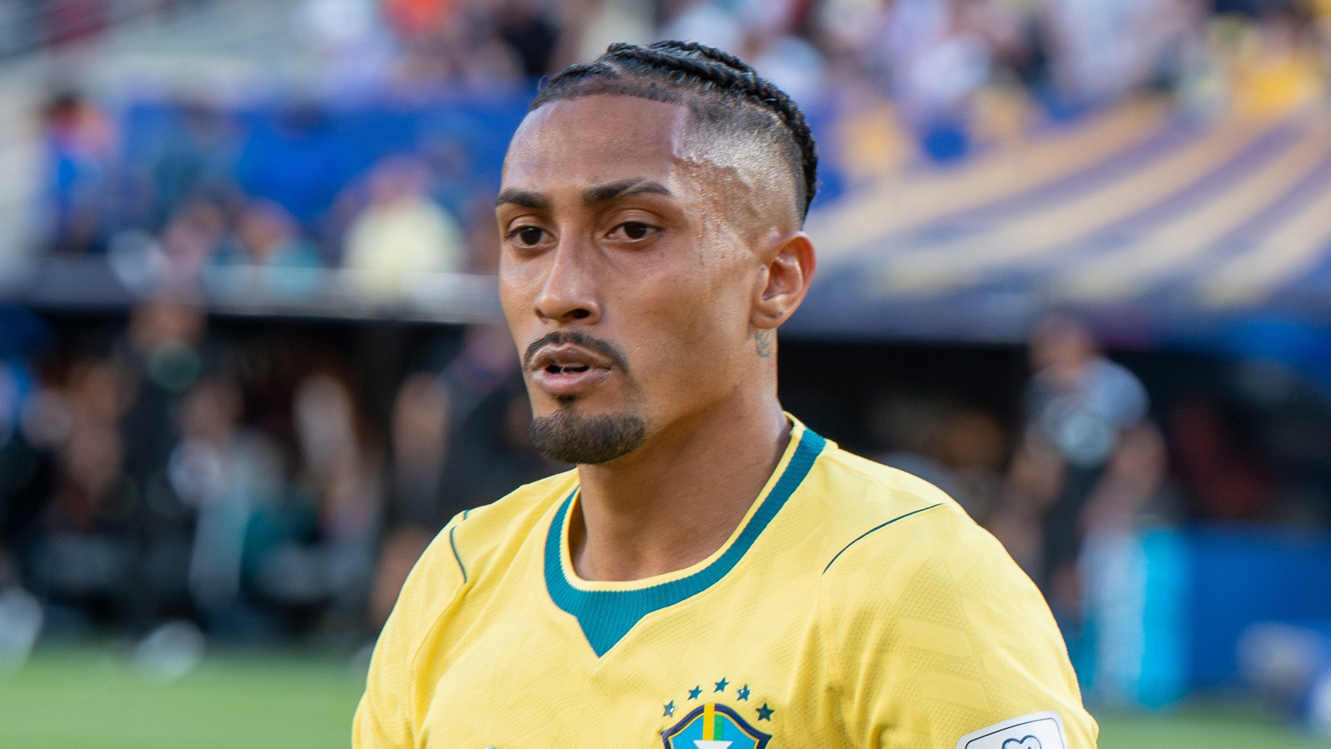 Close-up portrait of Raphinha in Brazil's yellow kit looking toward the left with focused expression.
